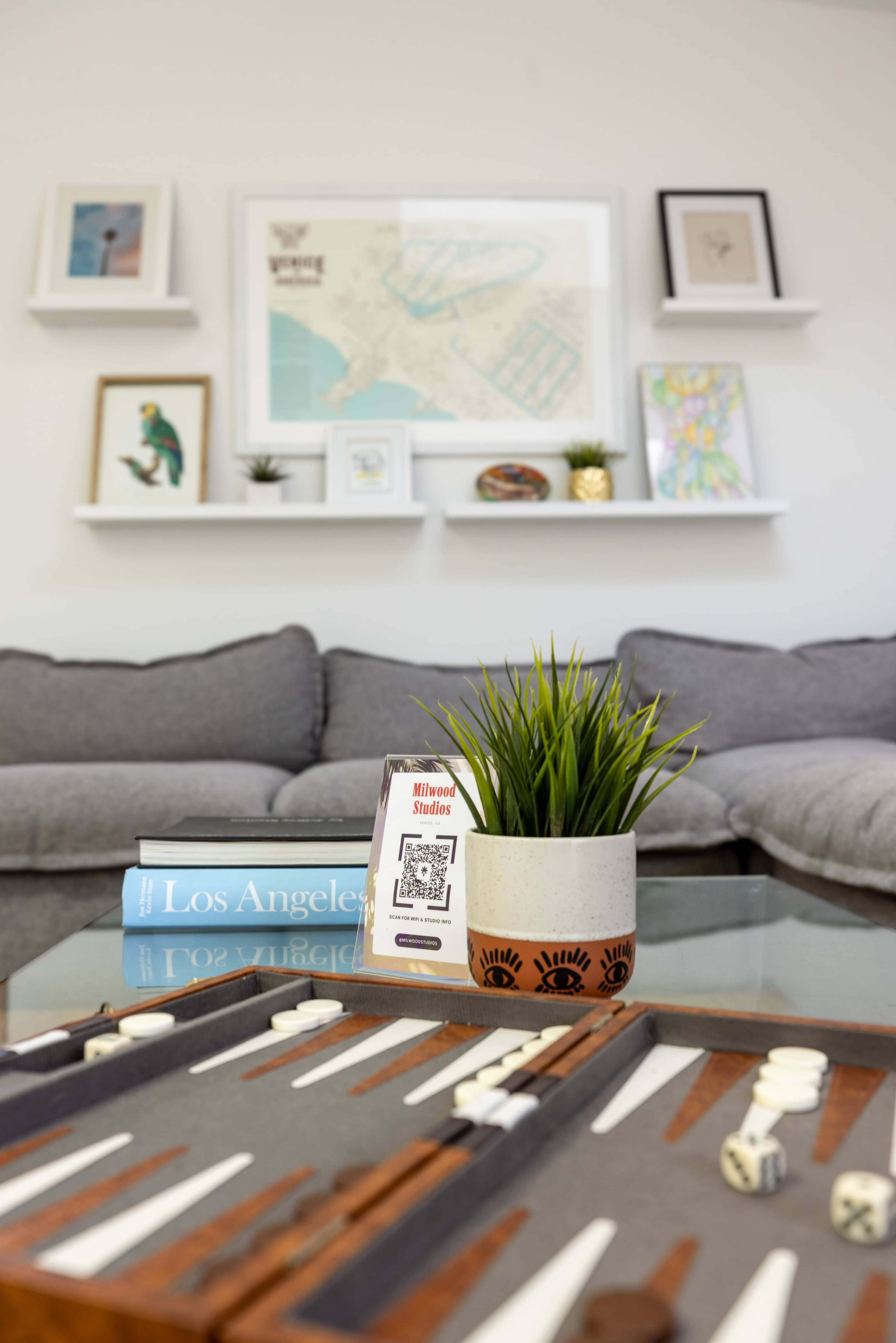 A coffee table with a backgammon set and a small potted plant sits in front of a couch in a stylish living room adorned with framed art.