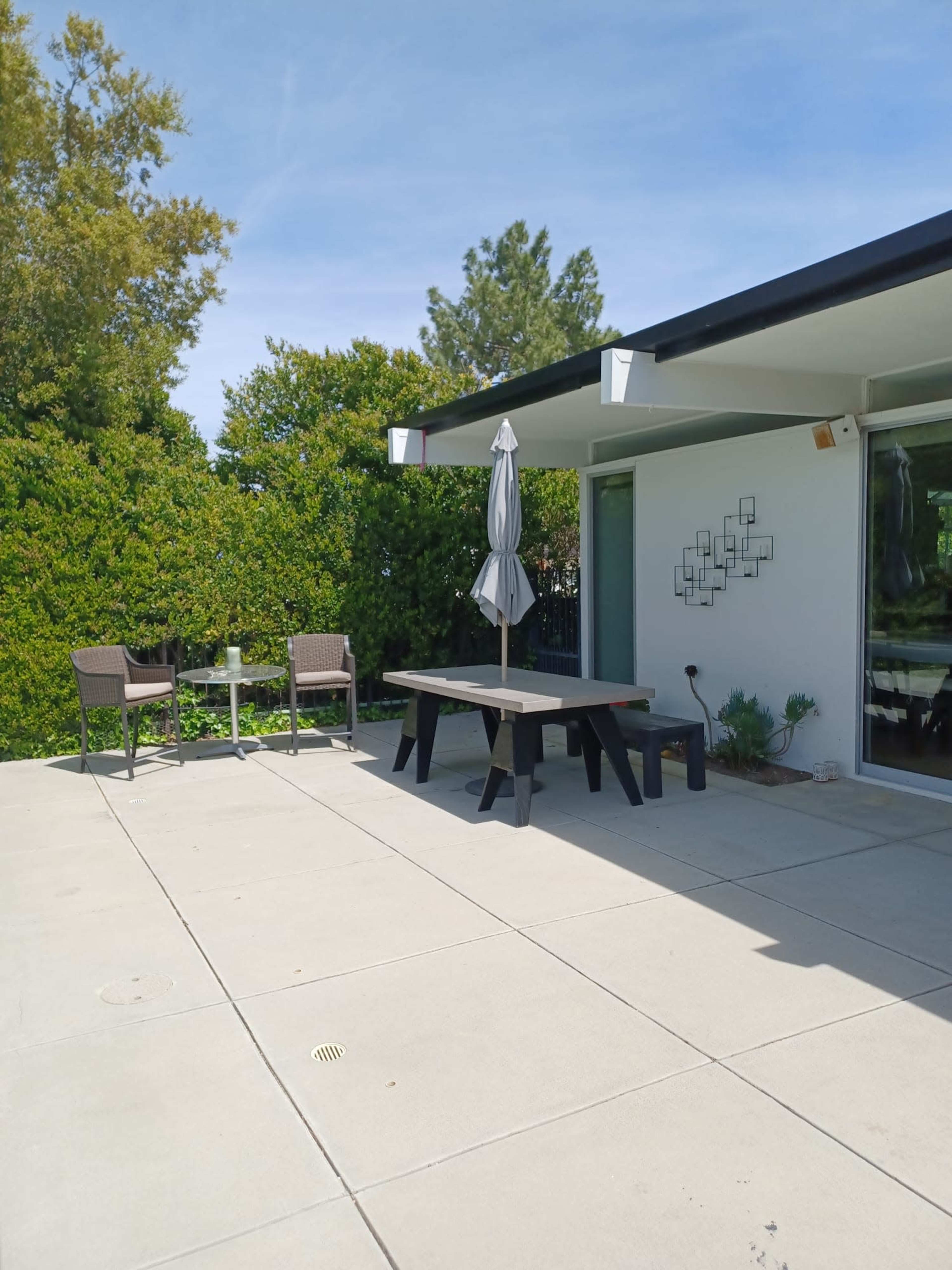 A modern patio features a large table with an umbrella, surrounded by two chairs, adjacent to a house and bordered by lush greenery.