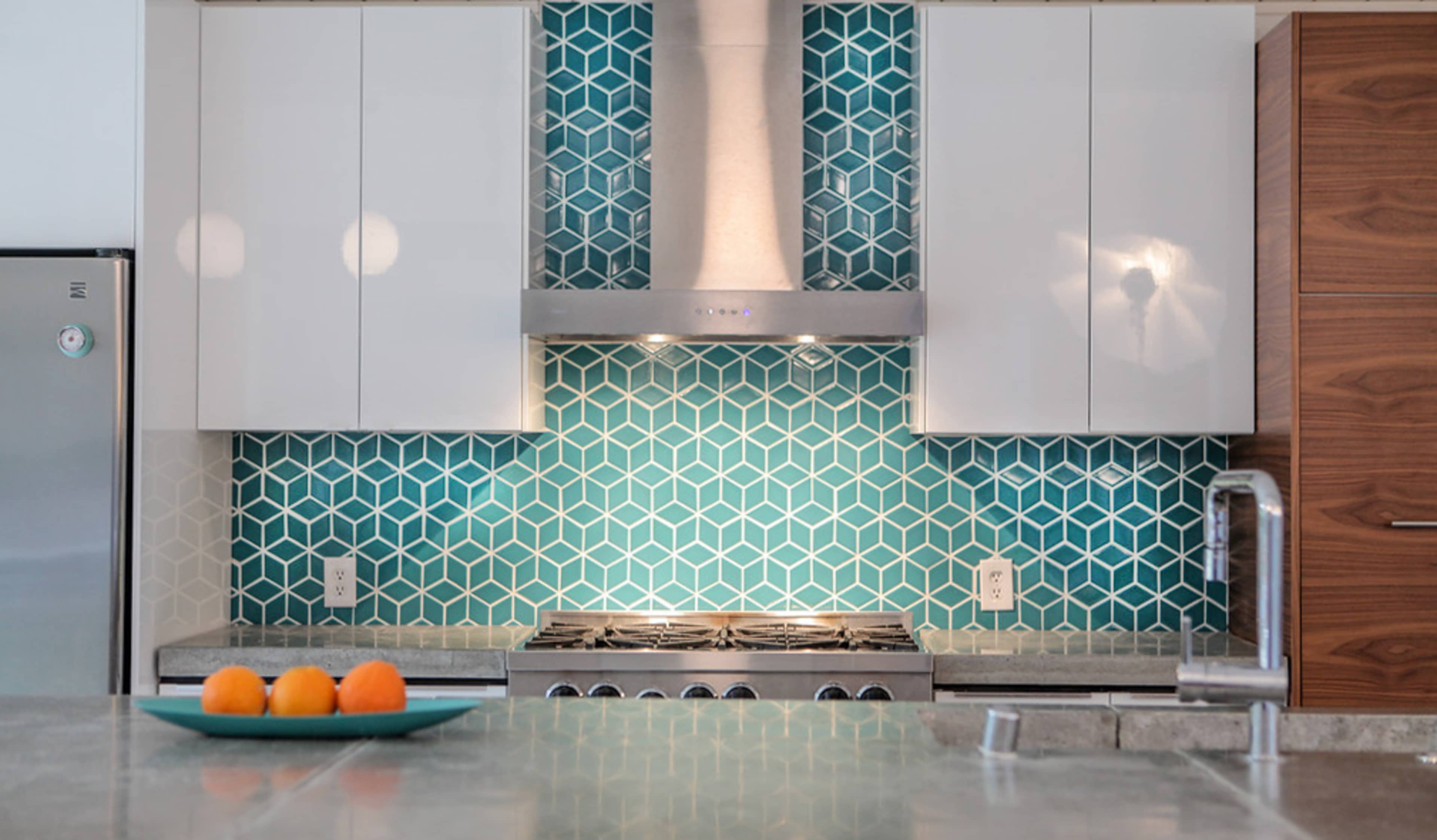 A modern kitchen features a geometric teal tile backsplash, stainless steel appliances, and a countertop with a bowl of oranges.