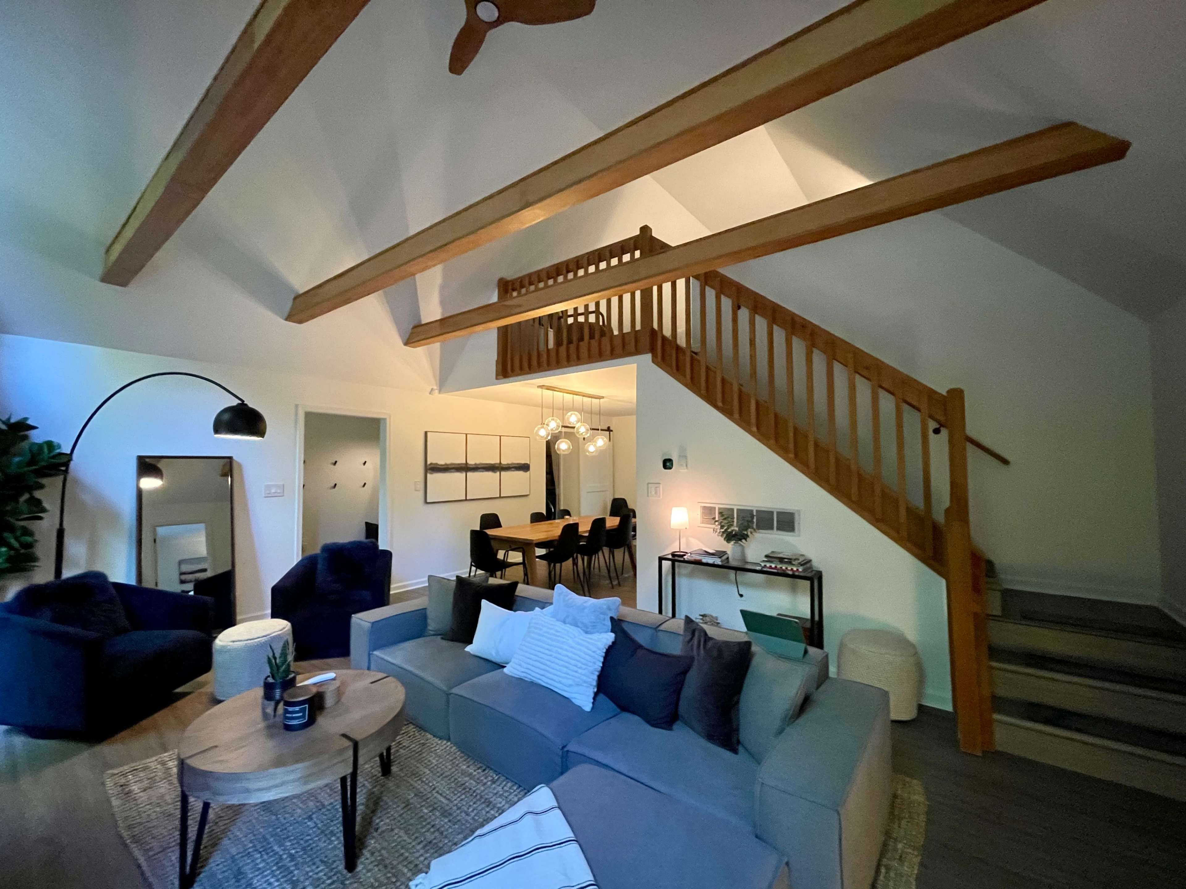 The image shows a spacious living area with a high ceiling, wooden beams, a large sectional sofa, a dining area in the background, and a staircase leading to an upper level.