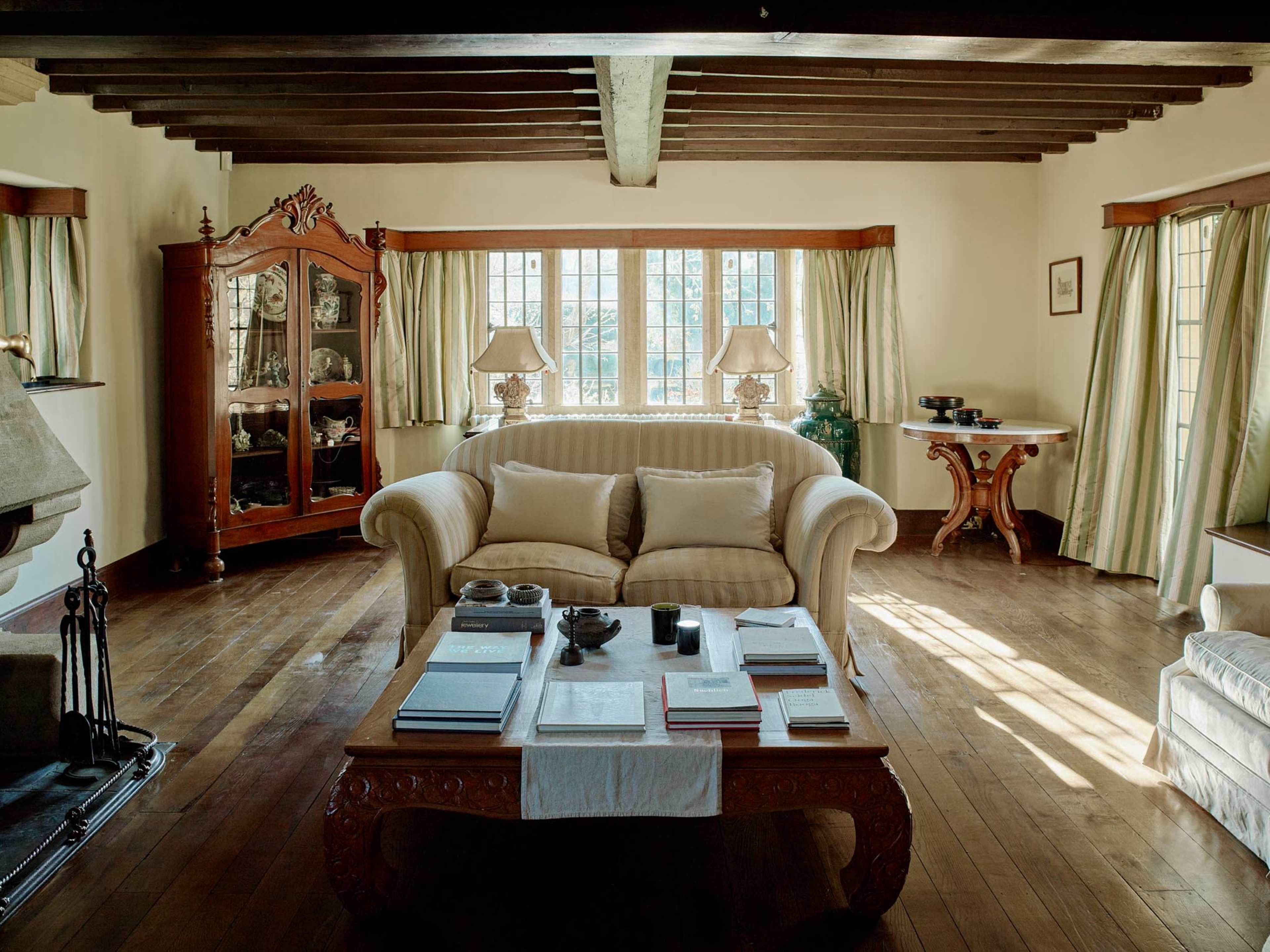 The image depicts a cozy living room featuring wooden beams, a plush beige sofa, a coffee table with various items, and large windows surrounded by light green curtains.
