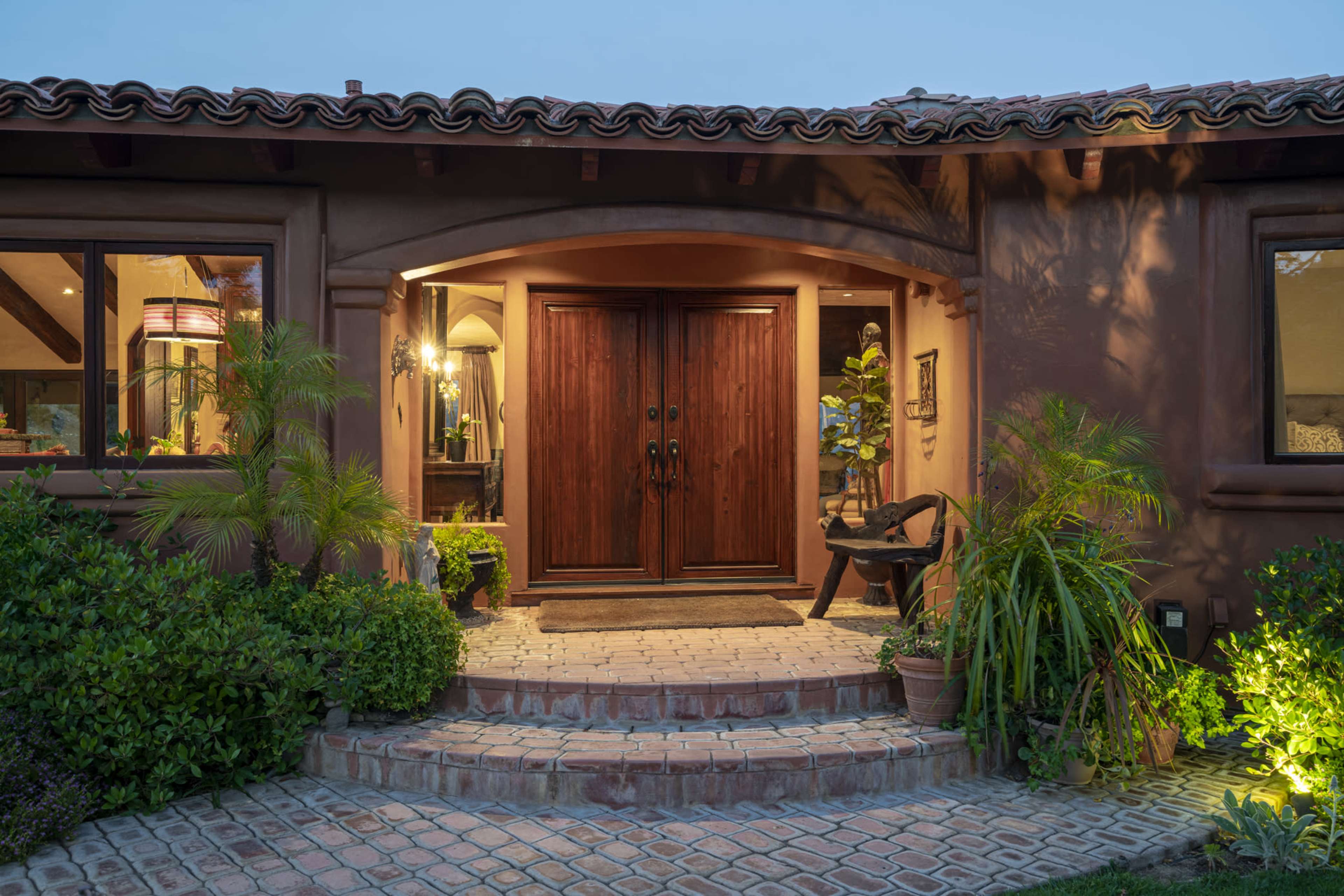 A warmly lit entrance features a set of double wooden doors flanked by potted plants and stone pathways leading to a cozy home.
