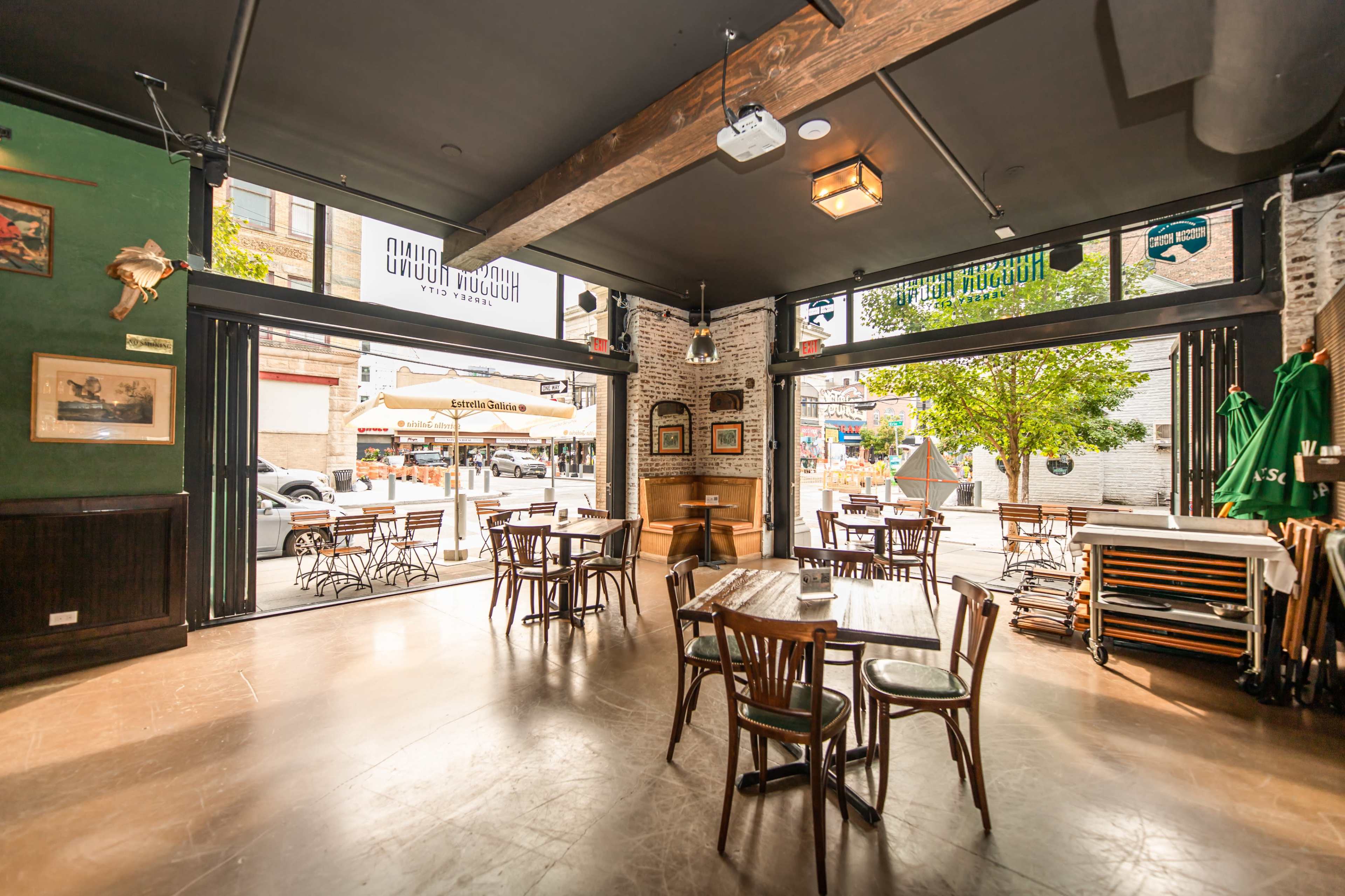 The interior of a restaurant features wooden tables and chairs, with large open windows facing a patio area.
