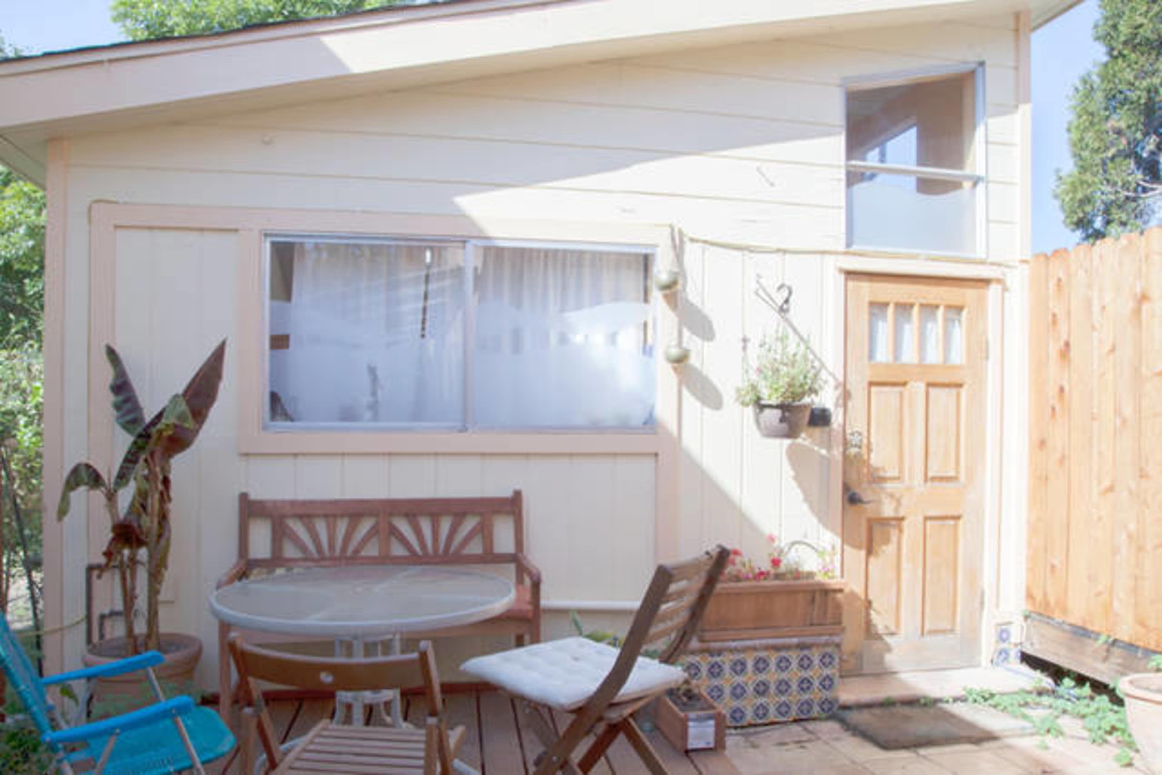 A small outdoor patio area in front of a light-colored house, featuring a wooden door, a round table with chairs, and potted plants.
