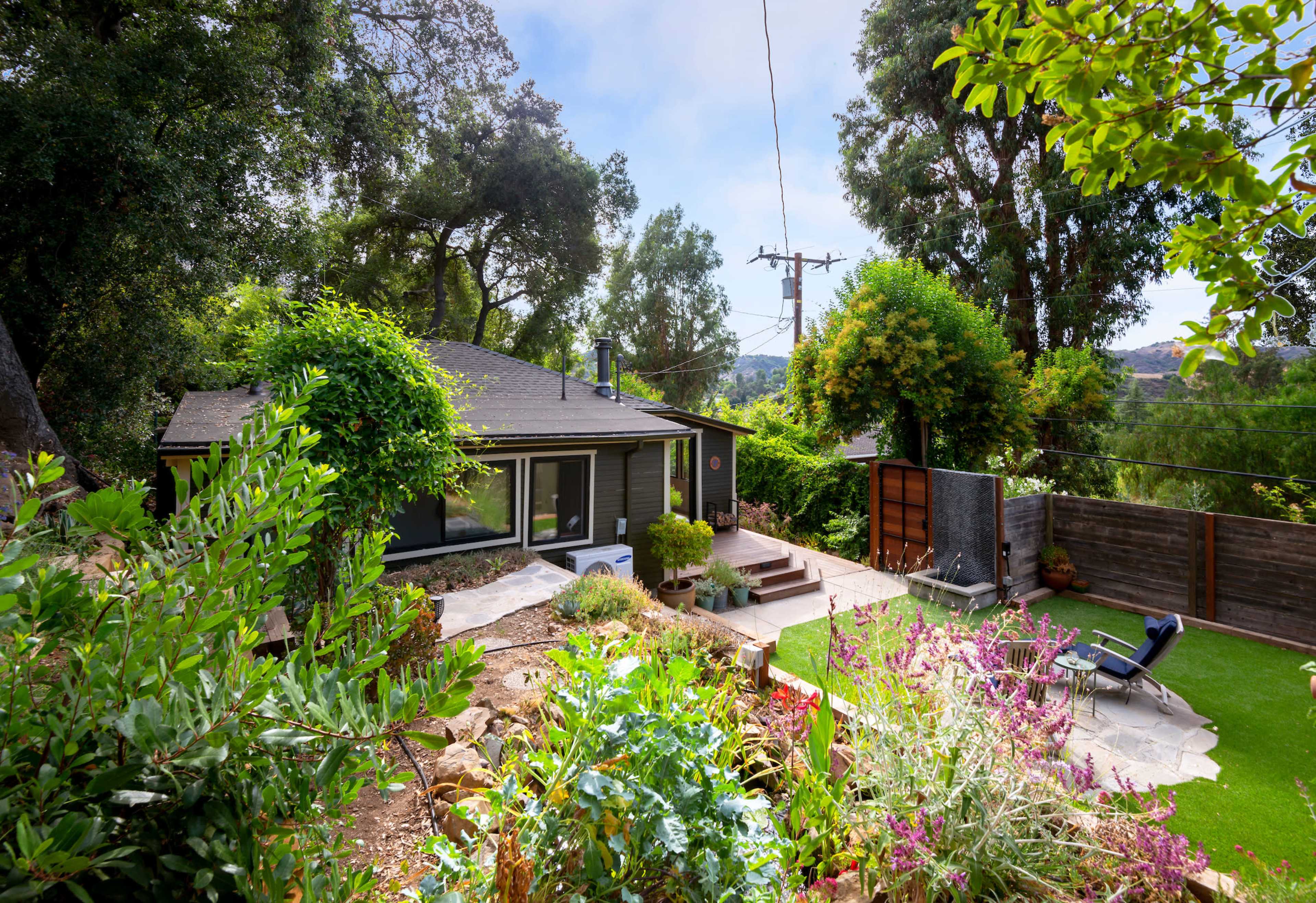 A small house is surrounded by lush greenery and colorful flowers, with a stone pathway leading to its entrance and a lawn area nearby.