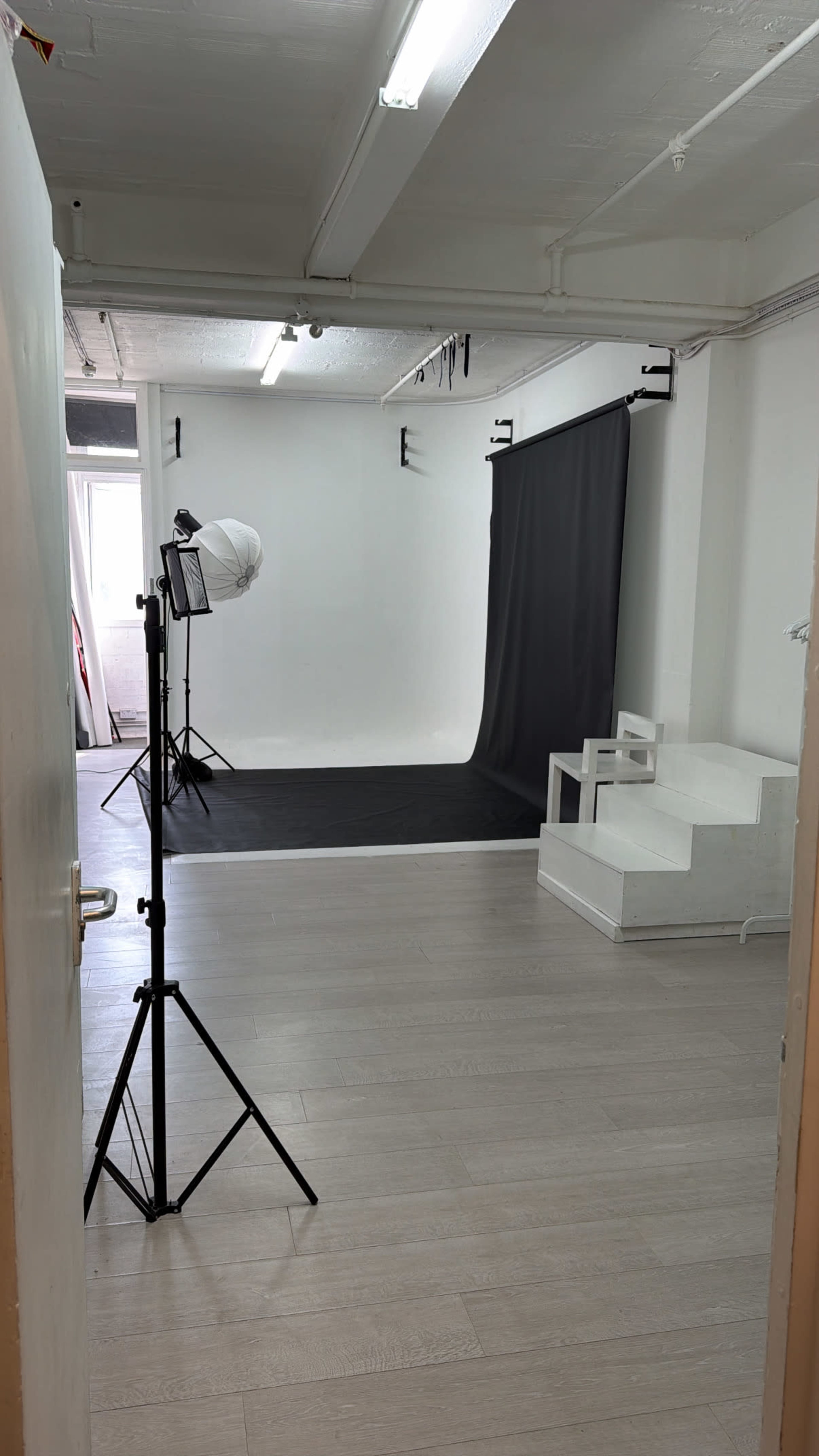 The image shows a photography studio with a black backdrop, a tripod light setup, and white steps in a well-lit room.