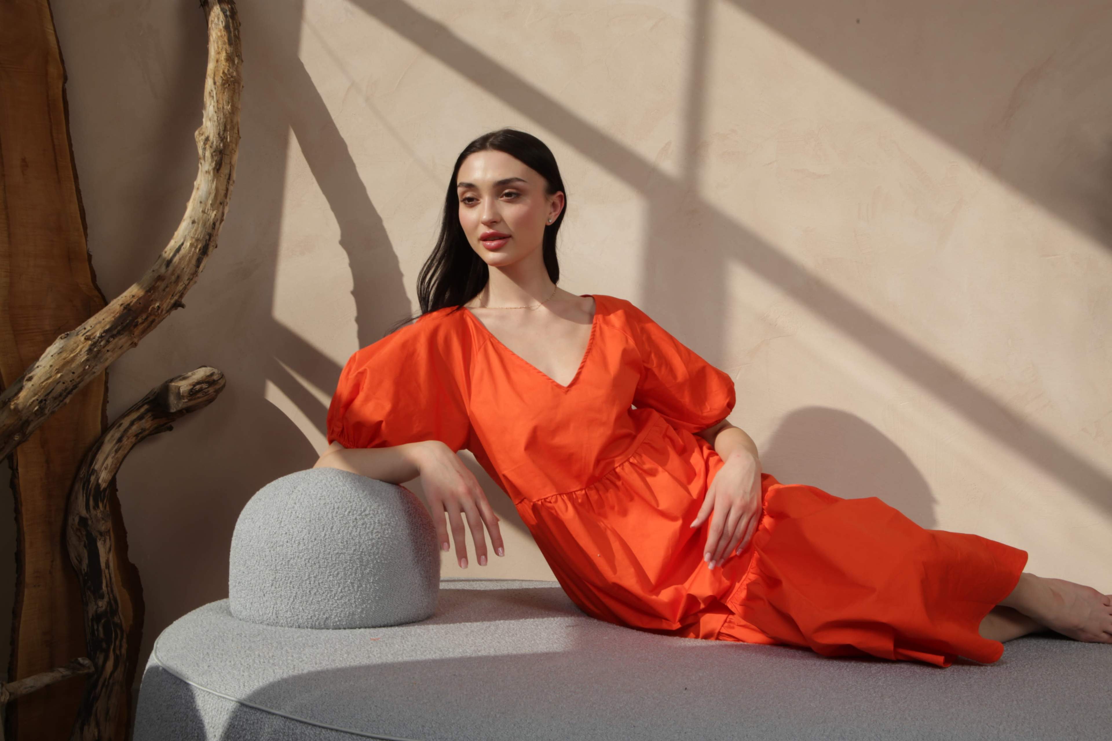 A woman in an orange dress poses on a gray, rounded sofa against a textured wall with natural light casting shadows.