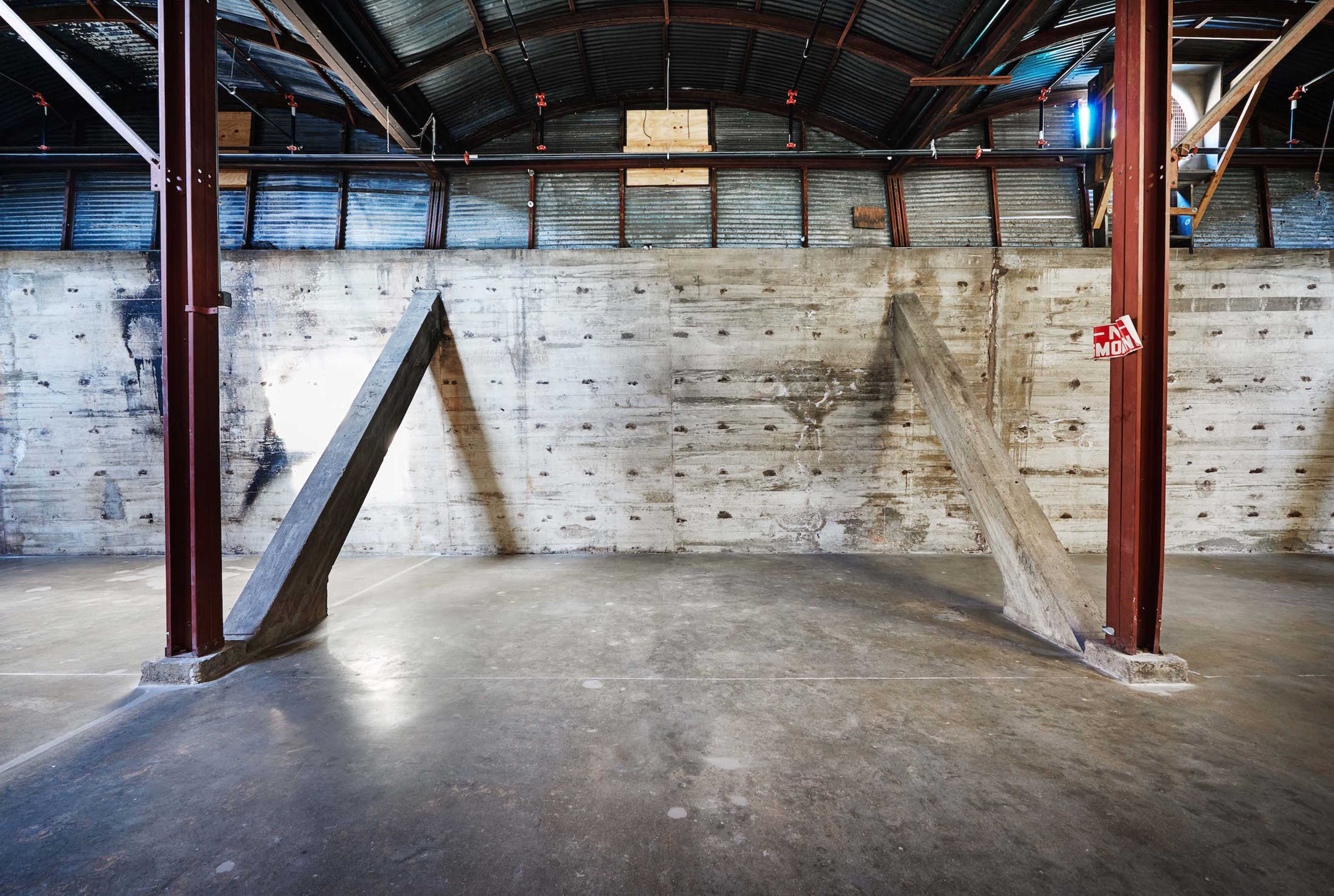 A large industrial space with exposed metal beams and concrete walls, featuring two diagonal concrete supports extending from the floor to the wall.