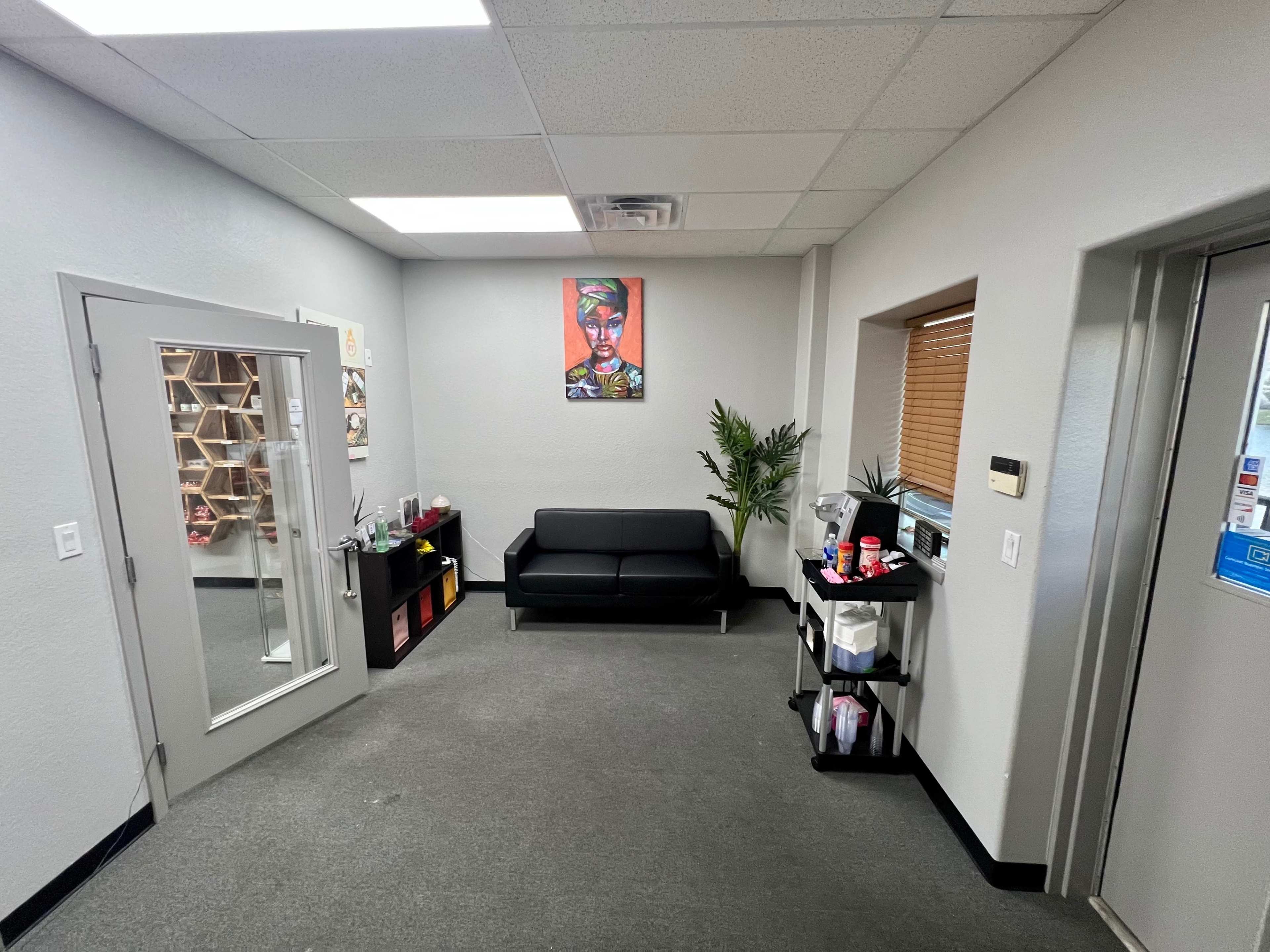 A waiting area with a black sofa, a potted plant, and artwork on the wall, along with a reception desk and shelves displaying items.