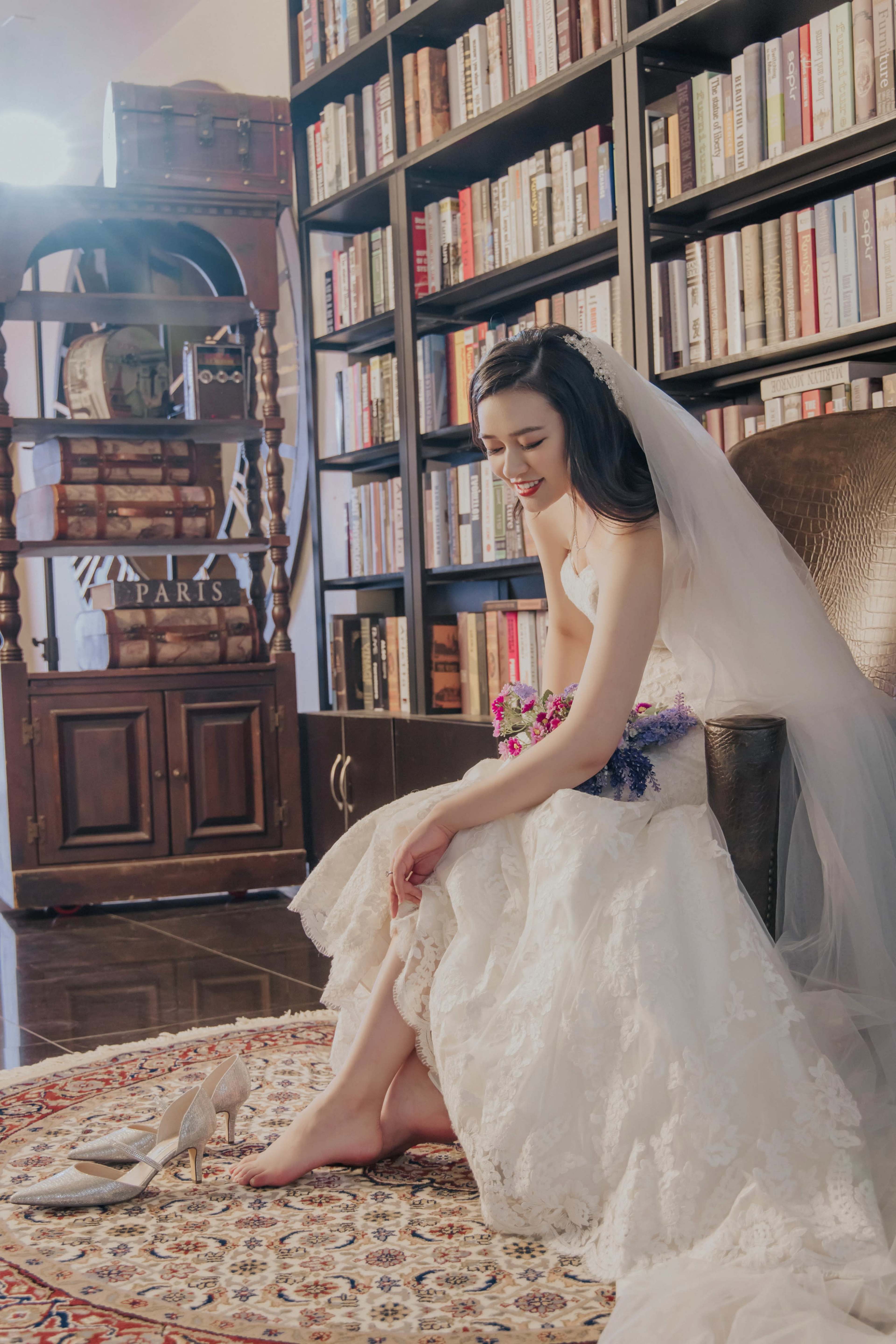 A bride in a wedding dress sits on a chair next to a bookshelf, smiling softly while placing her feet on a patterned rug.