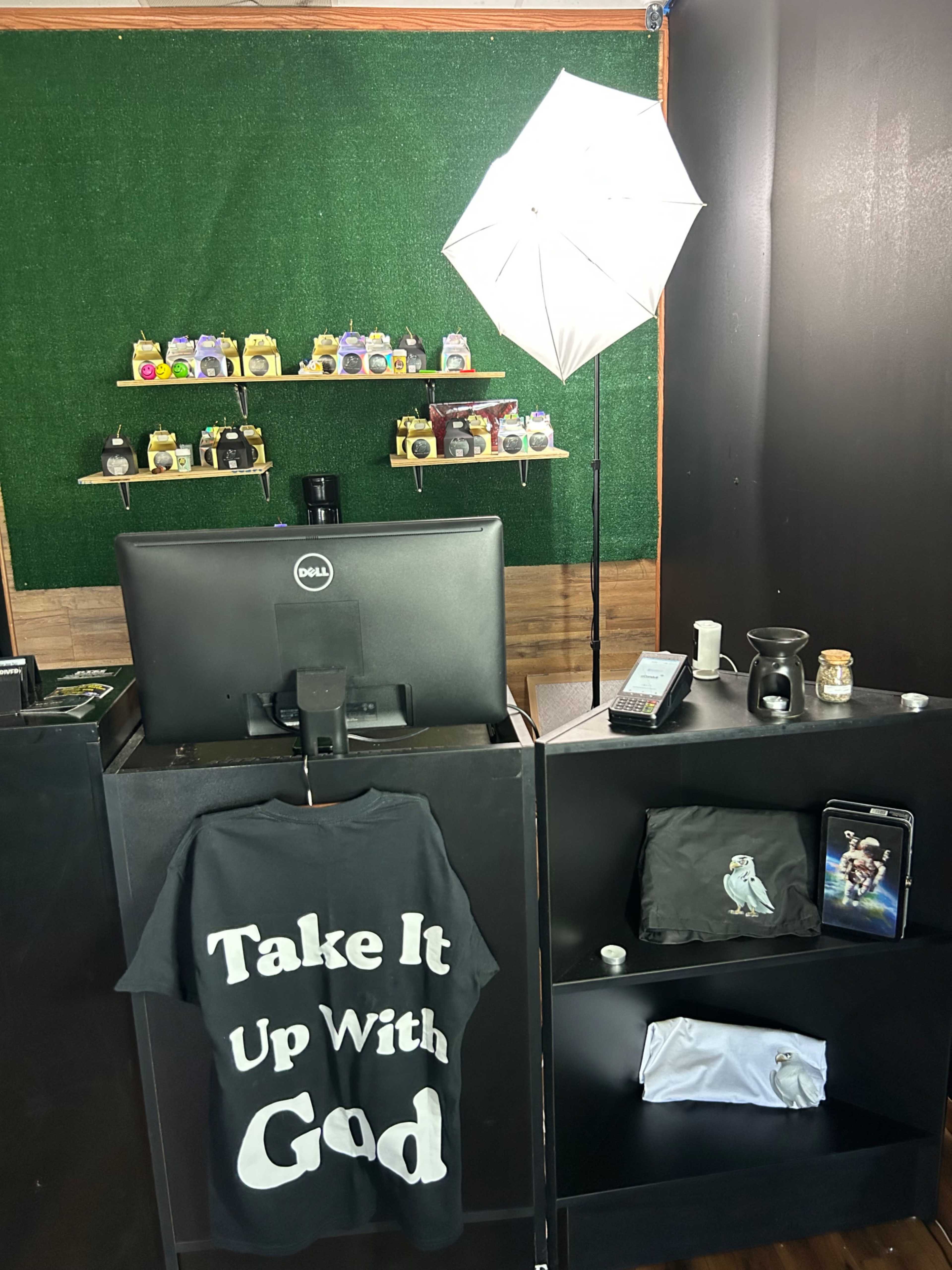 The image shows a retail counter featuring a computer monitor, a clothing rack with a shirt that reads "Take It Up With God," and various products displayed on shelves in the background.