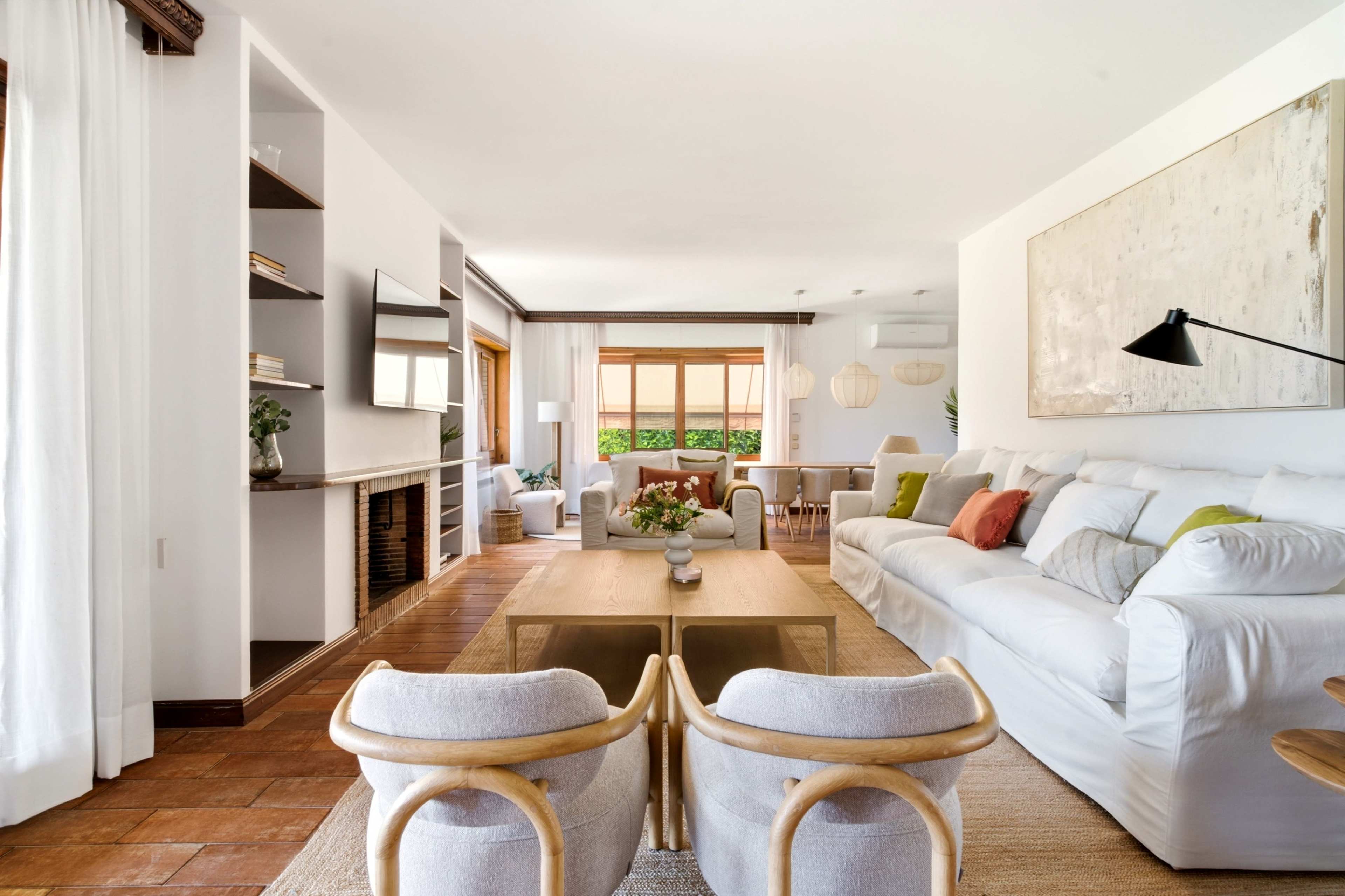 The image shows a modern living room with a white sectional sofa, a wooden coffee table, and a dining area visible in the background, featuring large windows and natural light.