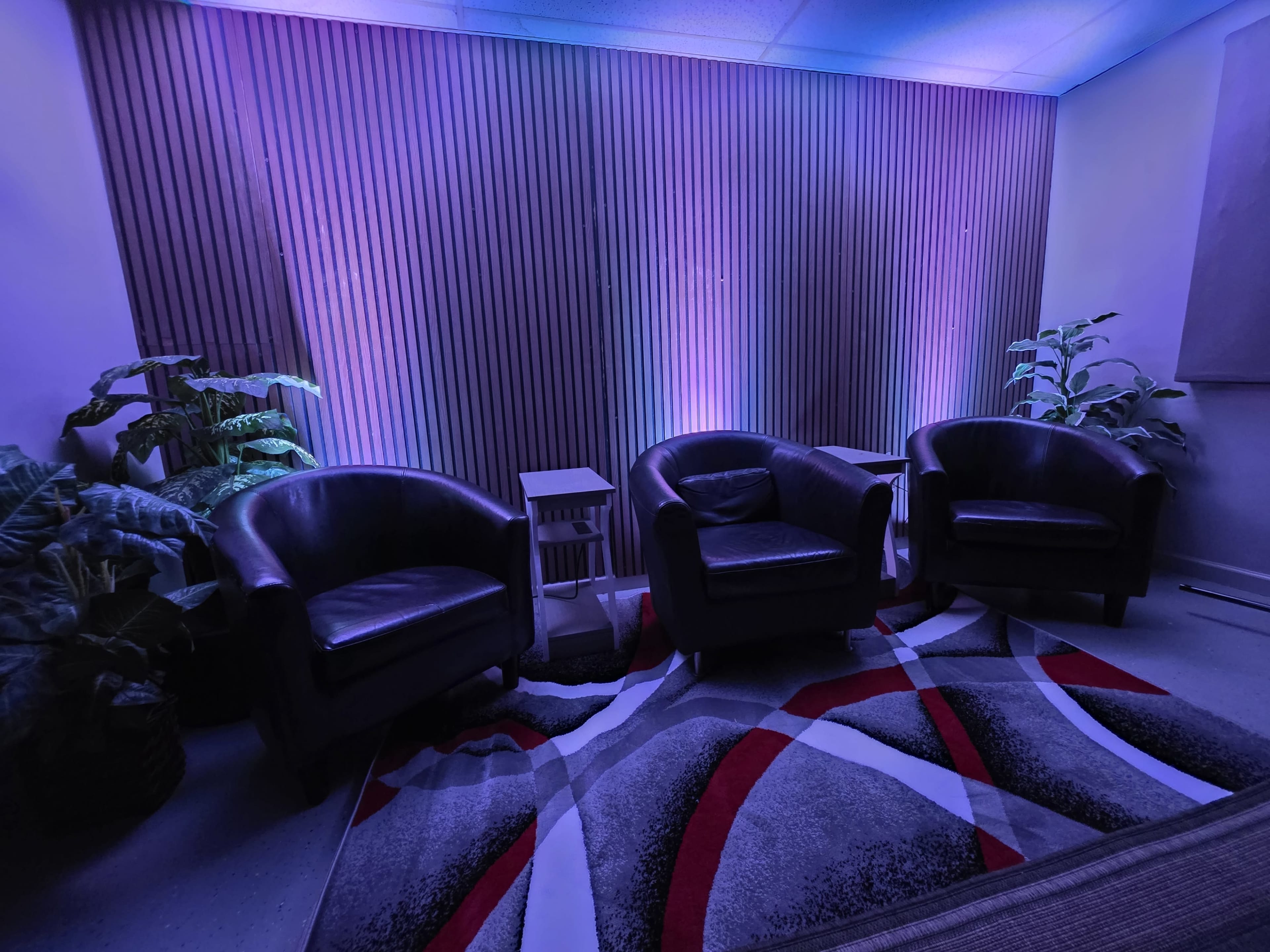 The image shows a cozy room with three black armchairs arranged around a modern rug, illuminated by purple and blue lighting against a wooden-paneled wall.