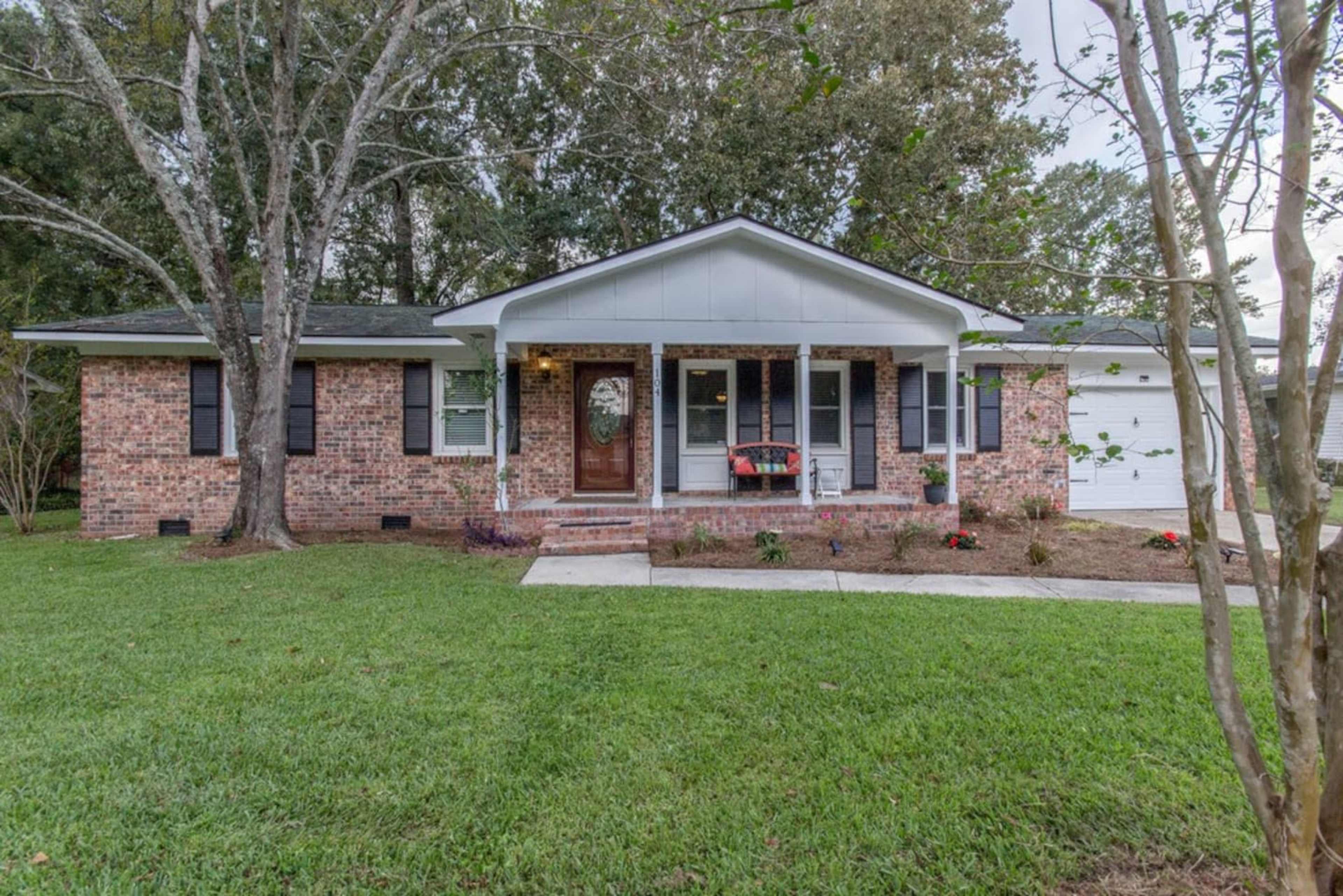 A brick house with a white roof and porch is surrounded by a green lawn and trees.