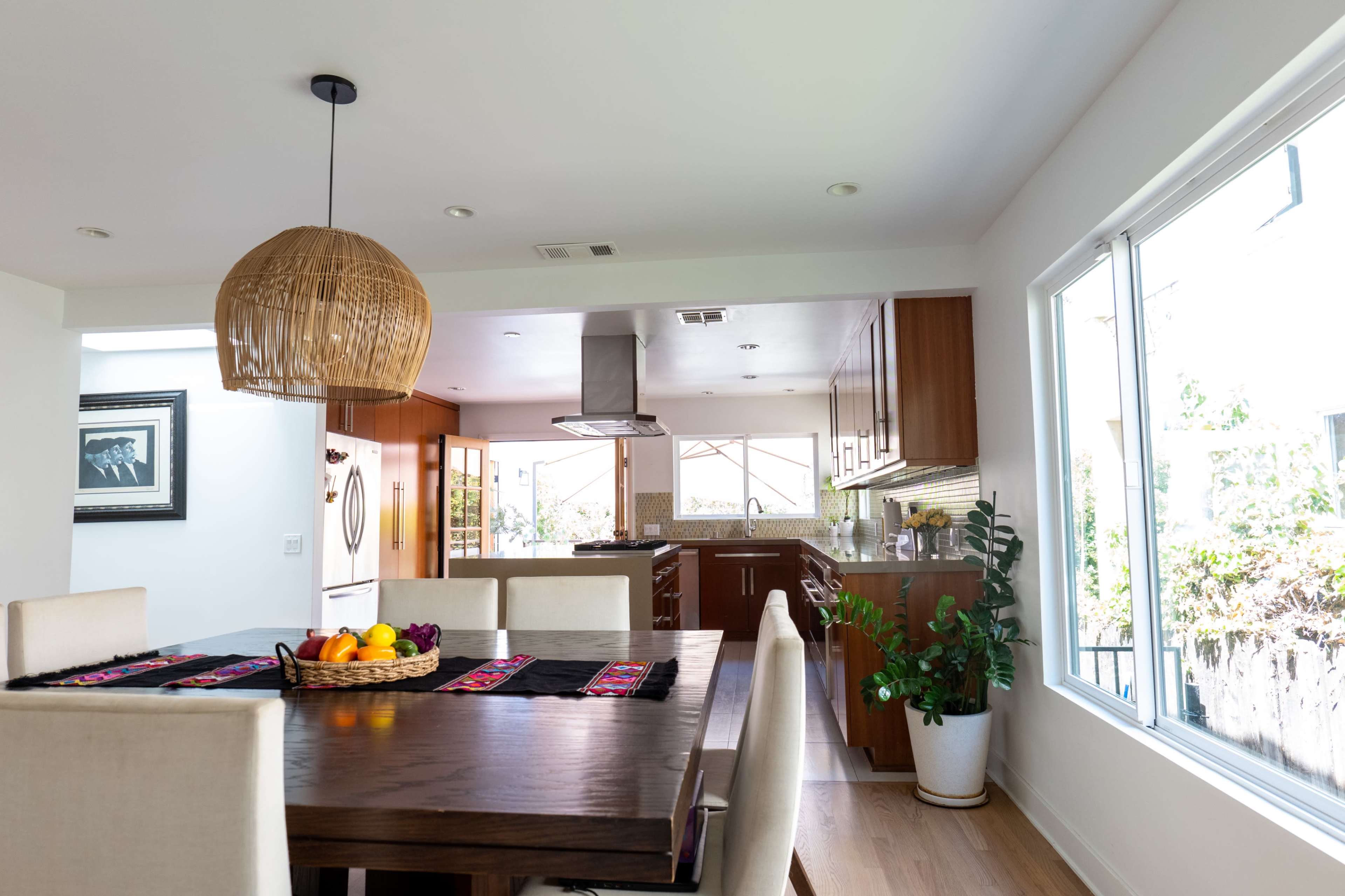 A spacious kitchen with wooden cabinets and a dining area featuring a large table, a woven pendant light, and a basket of fruit.