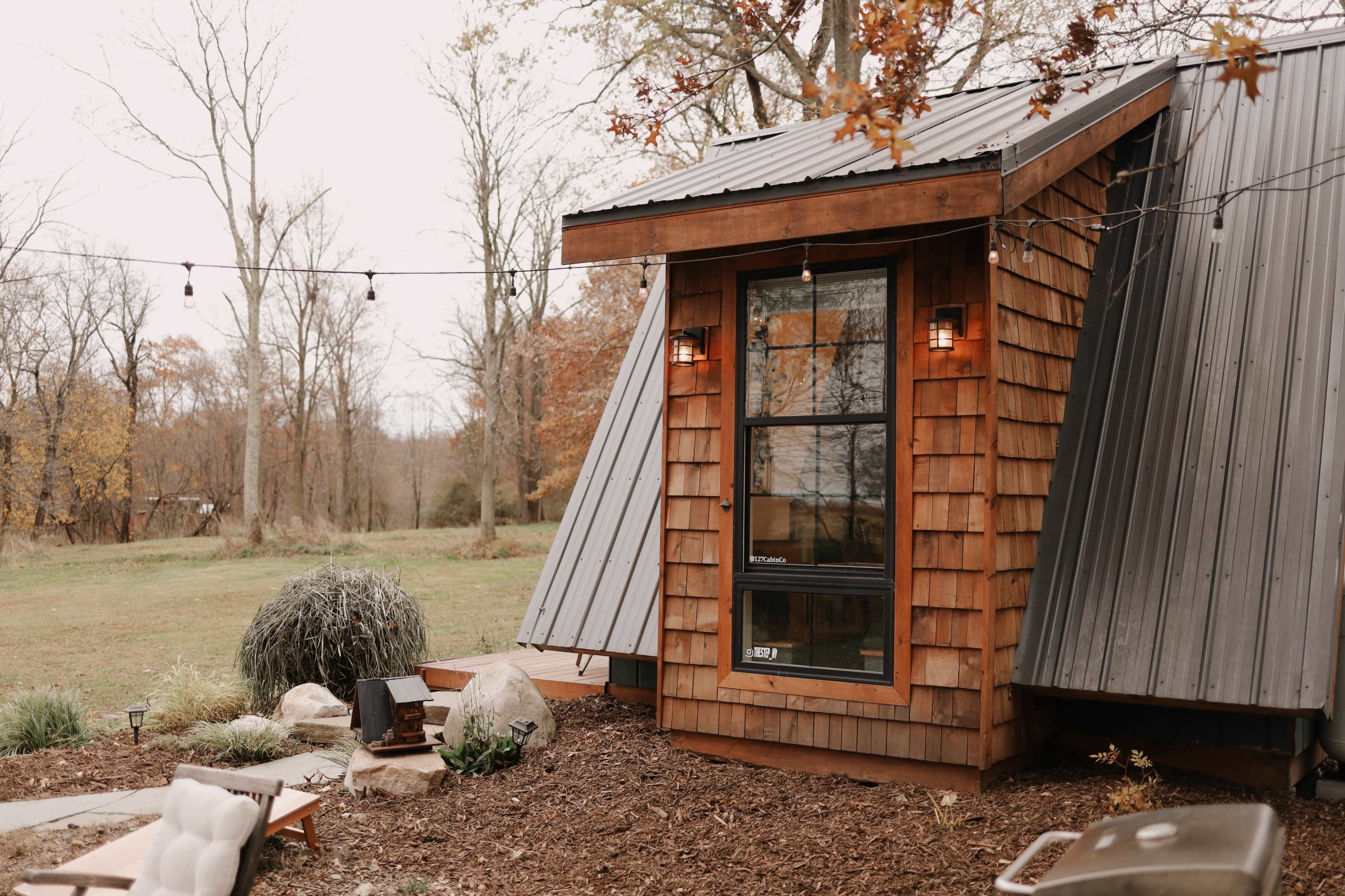 A modern wooden cabin with a sloped metal roof is situated in a grassy area surrounded by trees.