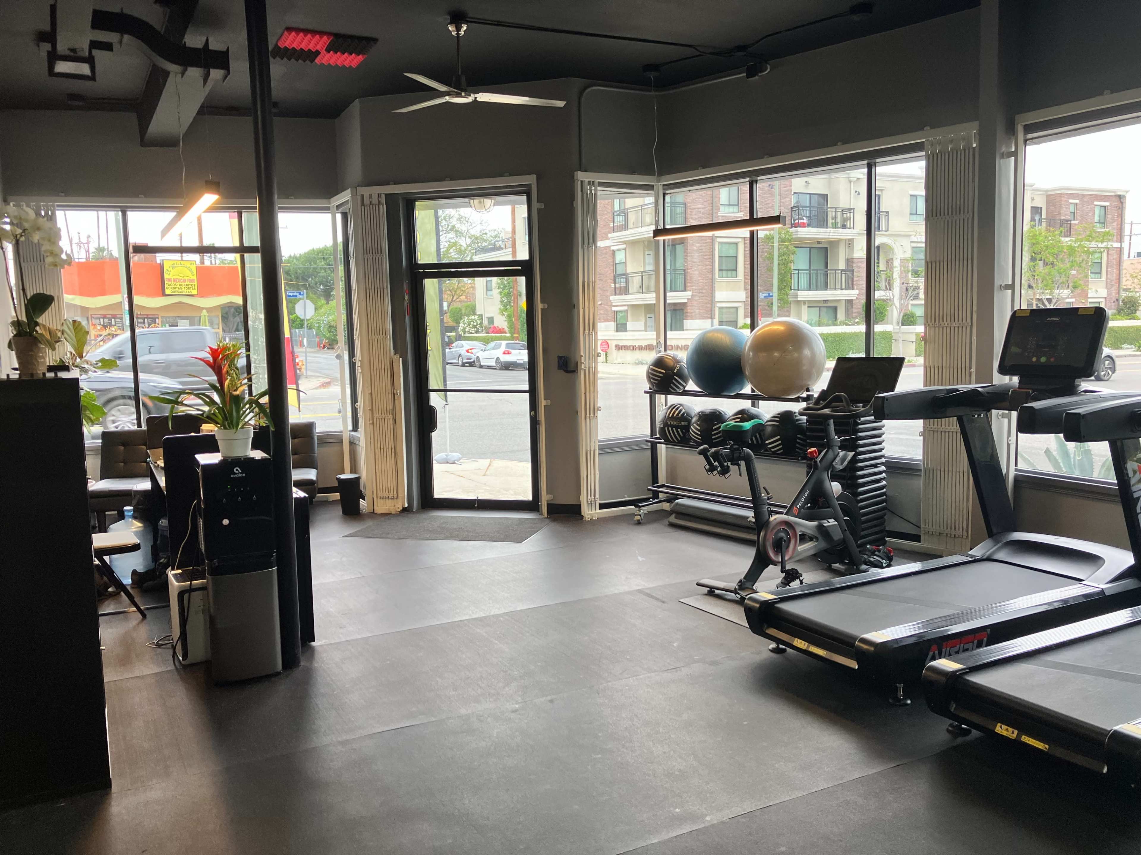 The image shows a gym interior with exercise equipment like treadmills and stability balls, large windows providing natural light, and a view of the street outside.
