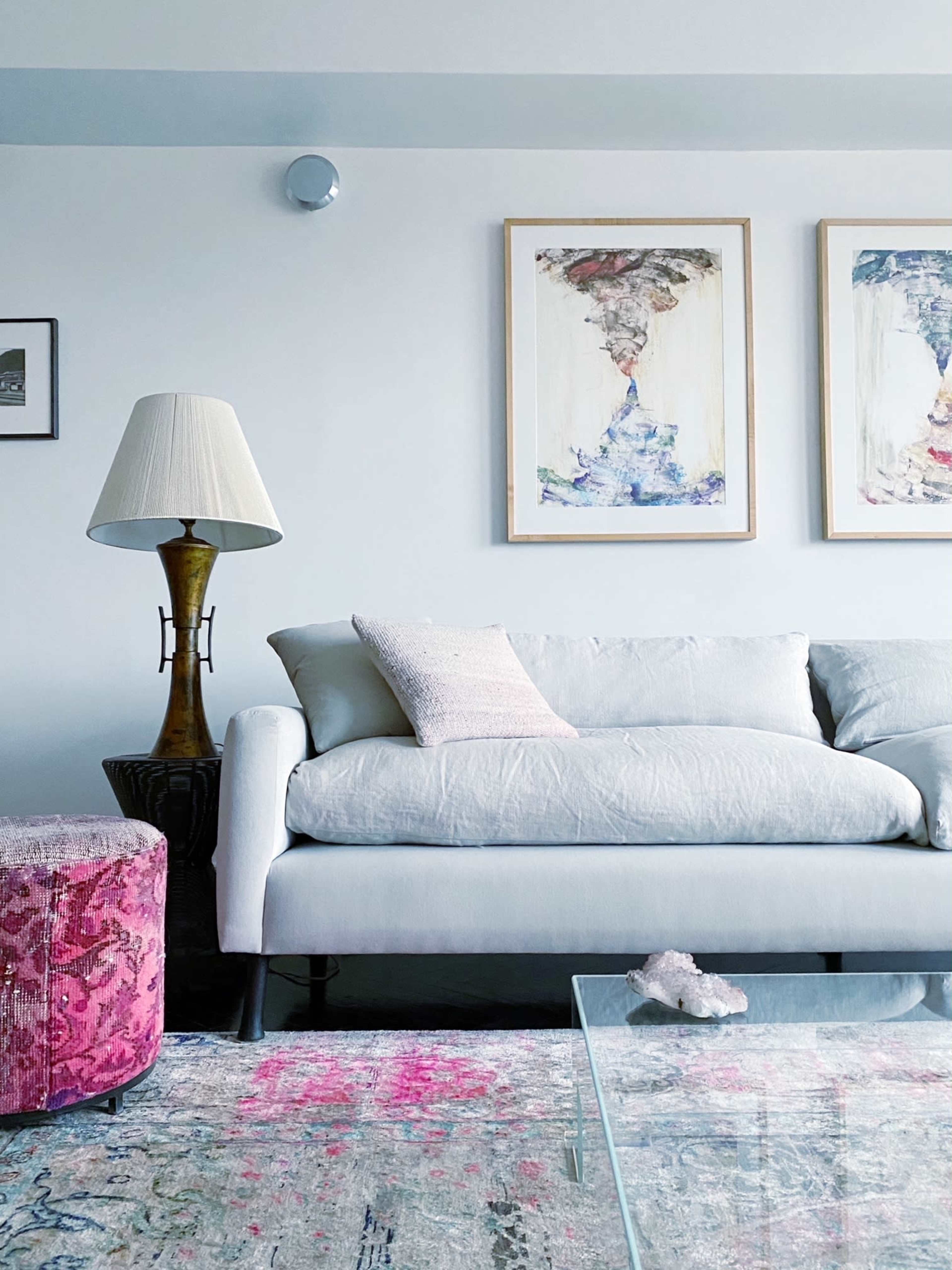 A light-colored couch with a decorative pillow, a lamp on a side table, and two framed artworks hanging on the wall above a colorful rug and a glass coffee table.