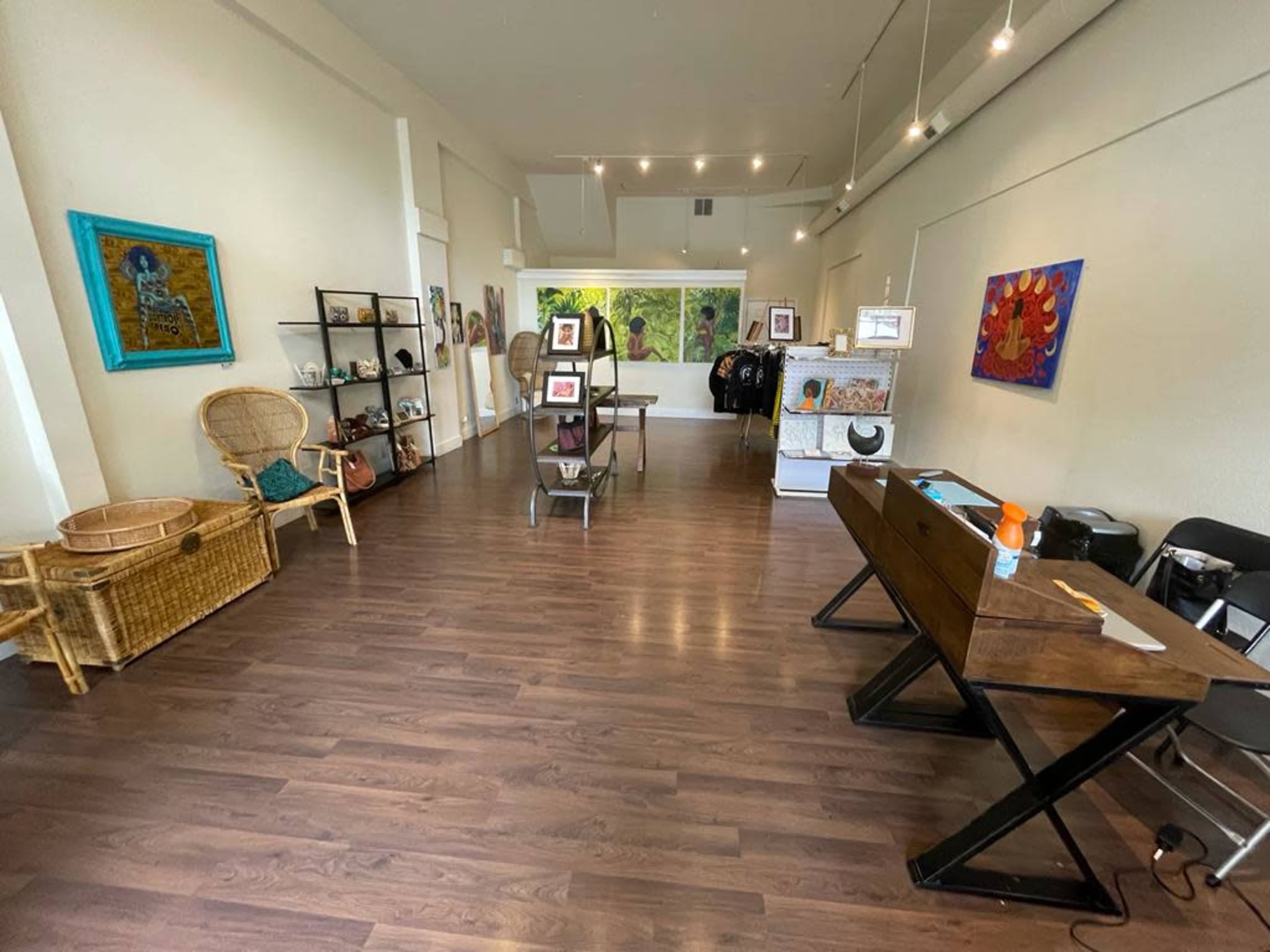 An interior of a retail space featuring wooden flooring, a few display tables, shelves with artworks, and large windows letting in natural light.