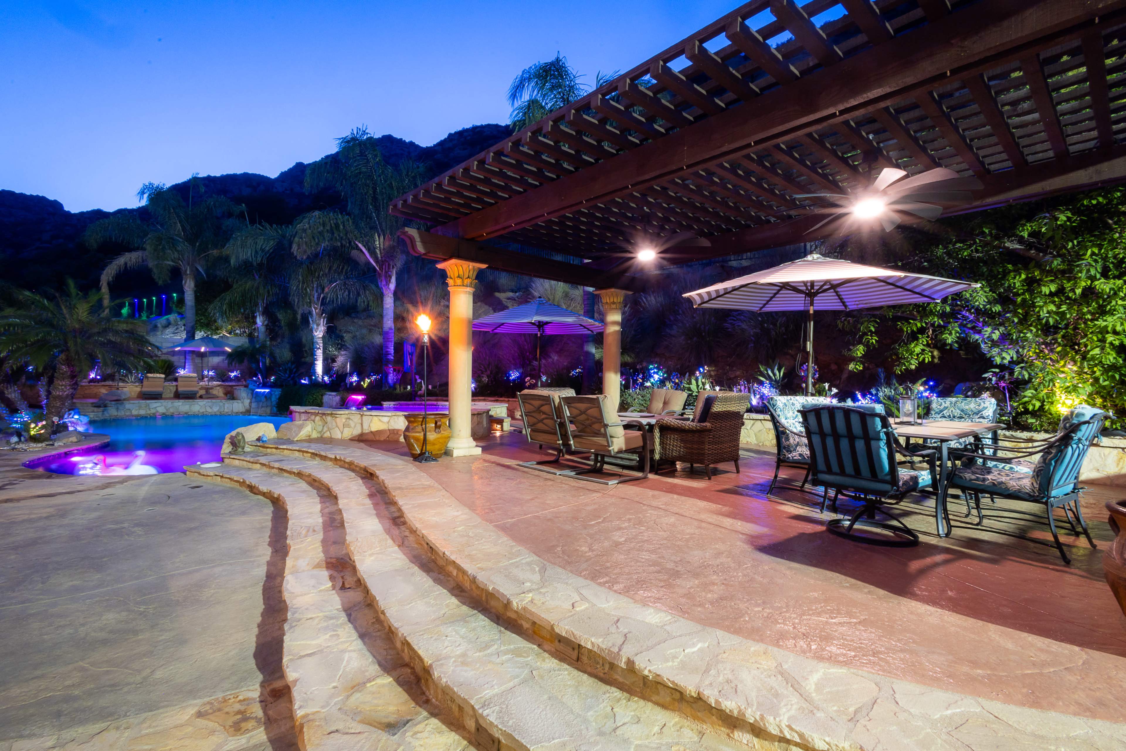 A landscaped outdoor space features a pool surrounded by palm trees, lounge chairs, and a shaded seating area under a pergola.