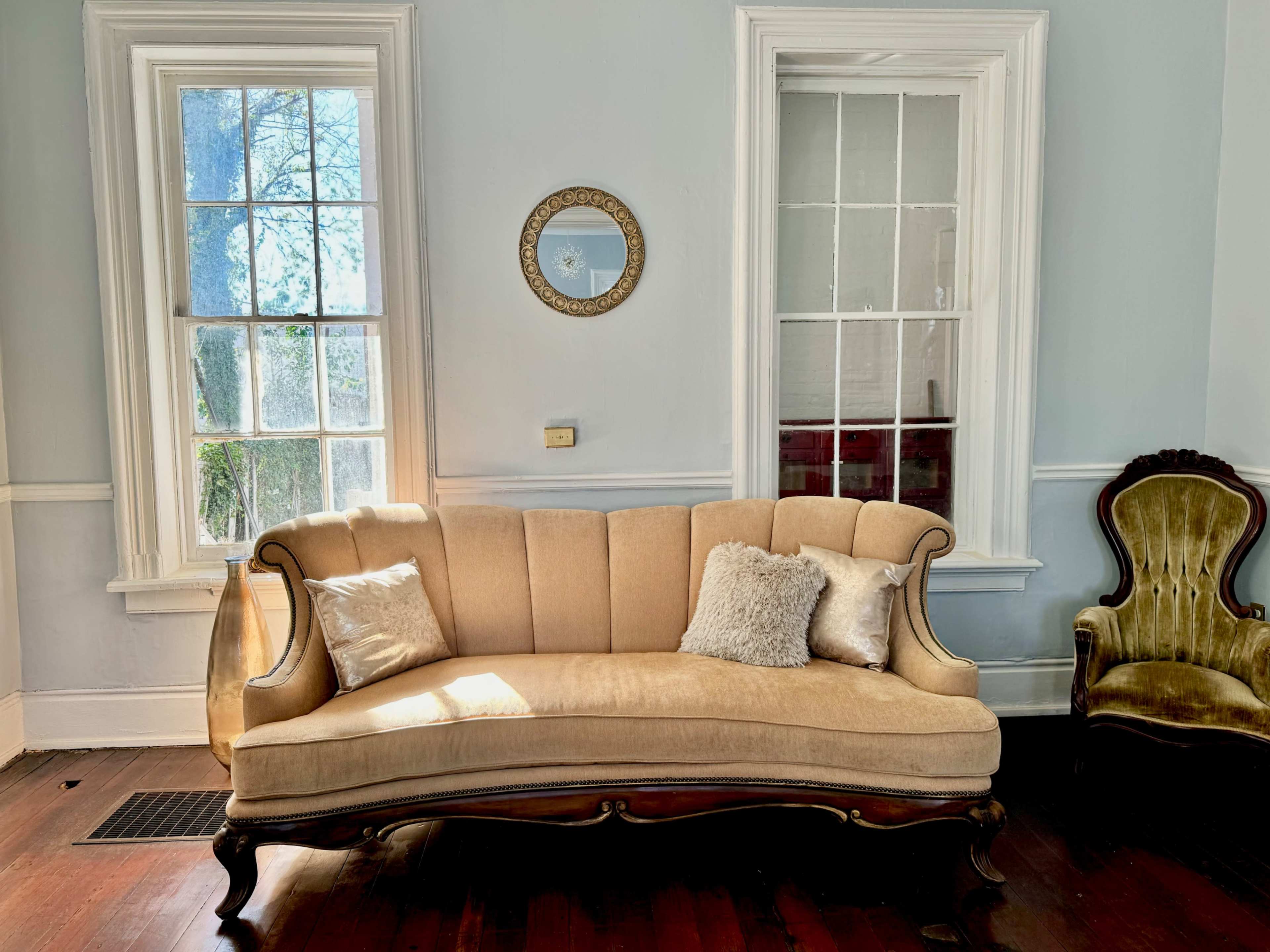 The image shows a beige upholstered couch against a wall with large windows, while a decorative mirror hangs above it and a vintage chair is positioned to the right.