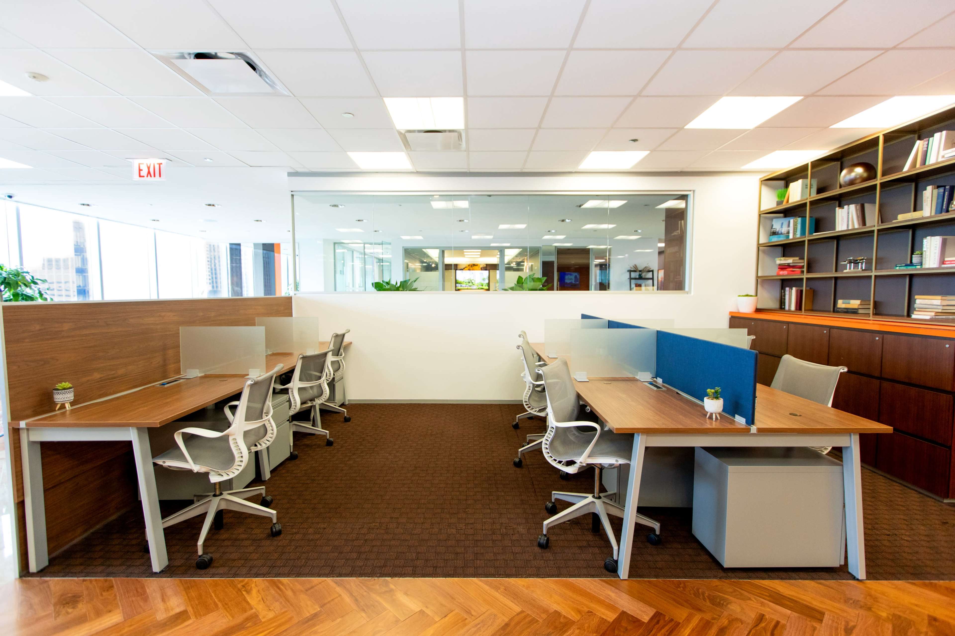 The image shows a modern office workspace with multiple desks arranged in a semi-enclosed layout, featuring ergonomic chairs and a large glass window separating it from another area.