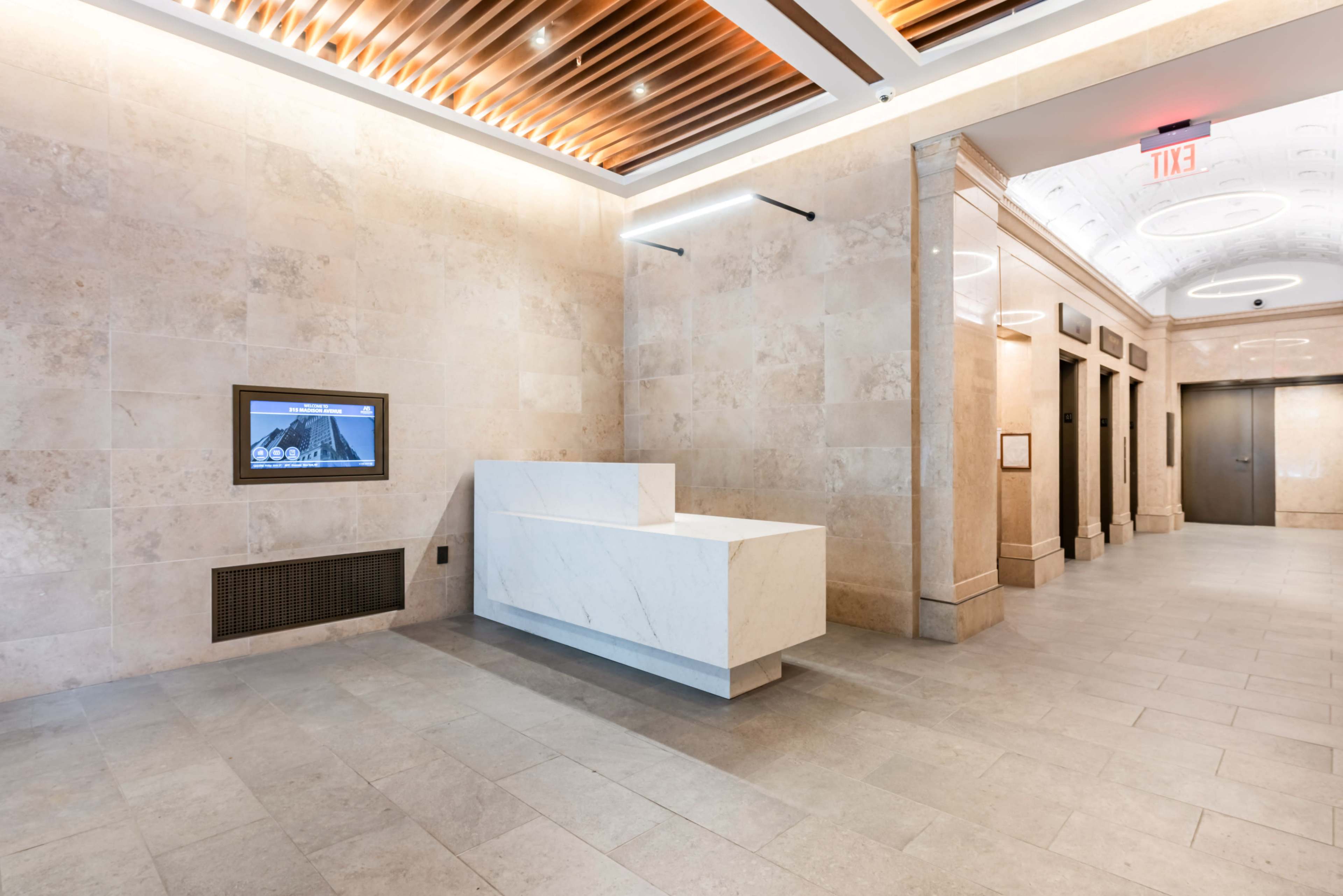 The image shows a modern lobby with a marble reception desk and elevators in a well-lit space featuring beige stone walls and a wooden ceiling.