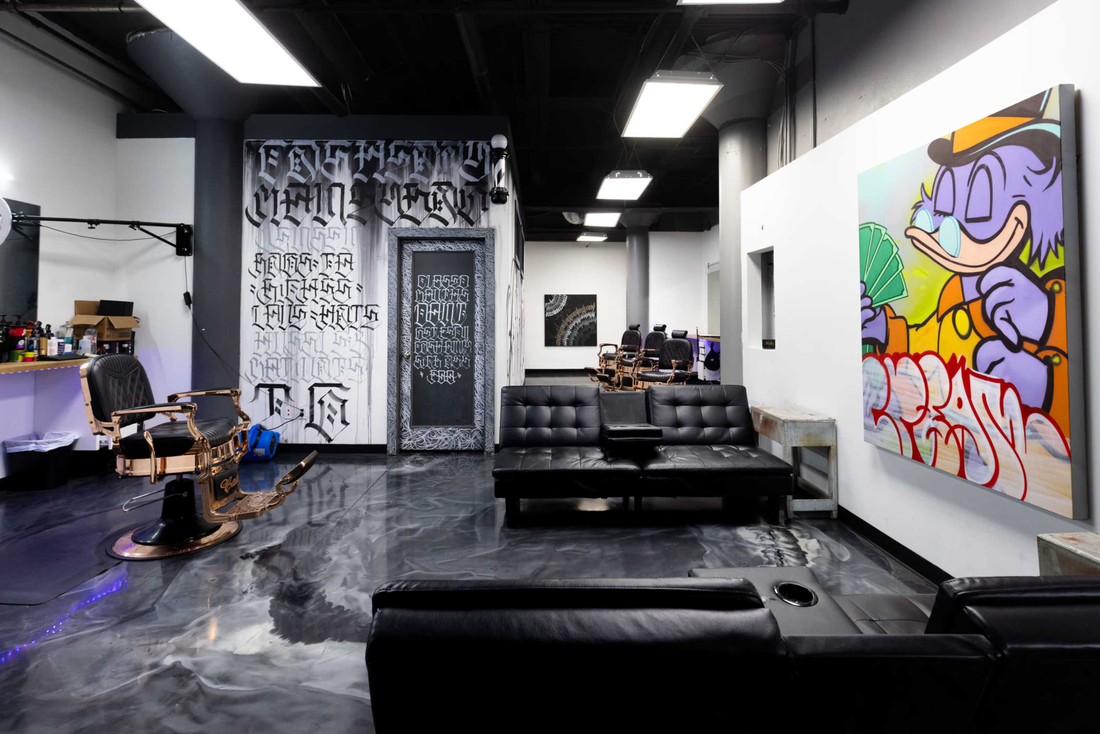 The image shows a stylish barbershop interior featuring modern black furniture, artistic wall murals, and an industrial design aesthetic.