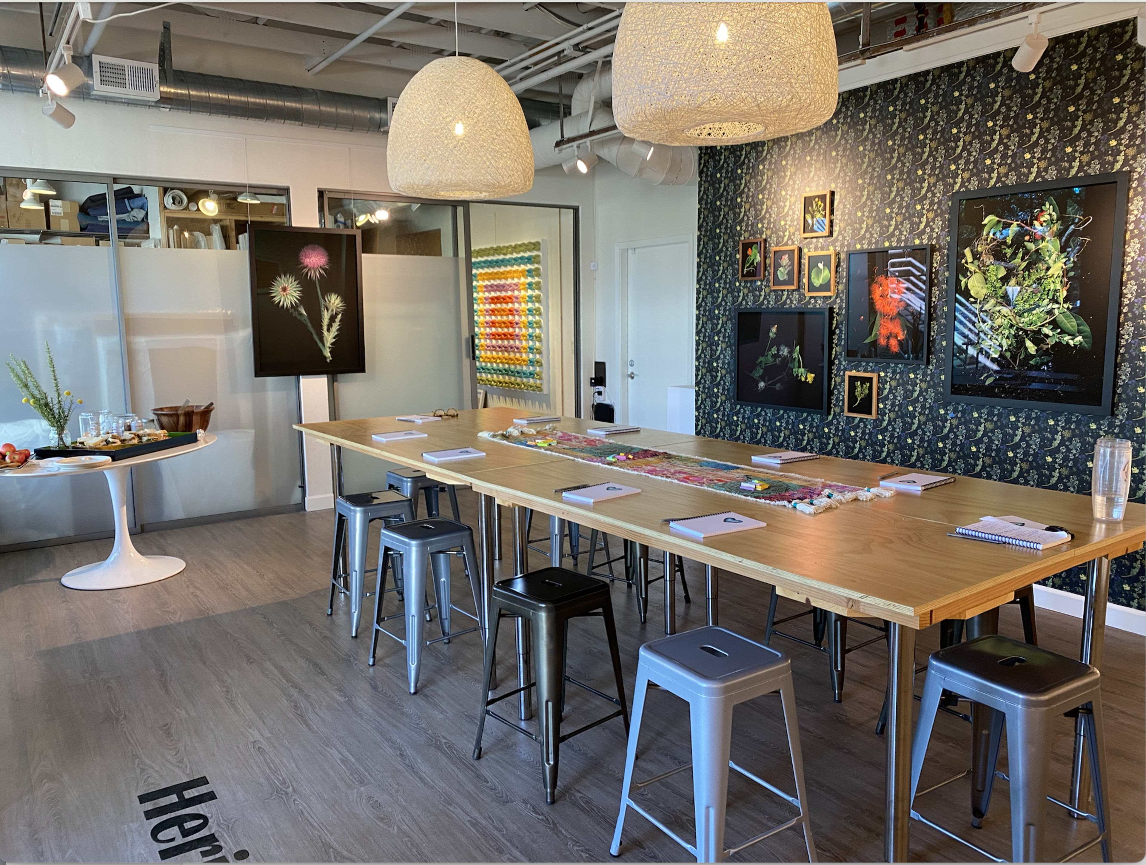 A modern dining or meeting space with a long wooden table surrounded by metal stools, decorated walls, and framed artworks displayed.
