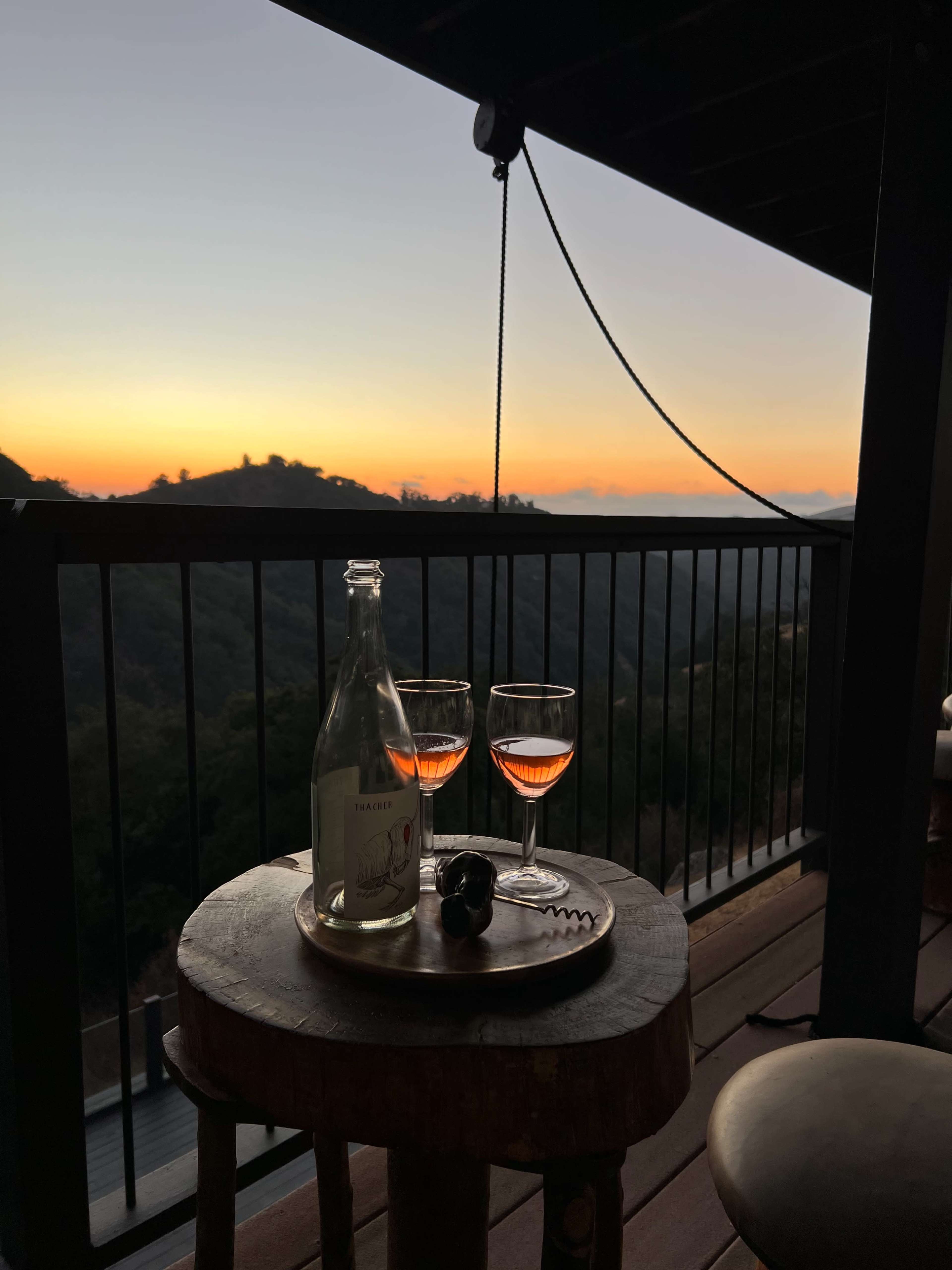 A bottle of sparkling wine and two glasses sit on a wooden tray atop a stool, with a sunset landscape visible in the background.