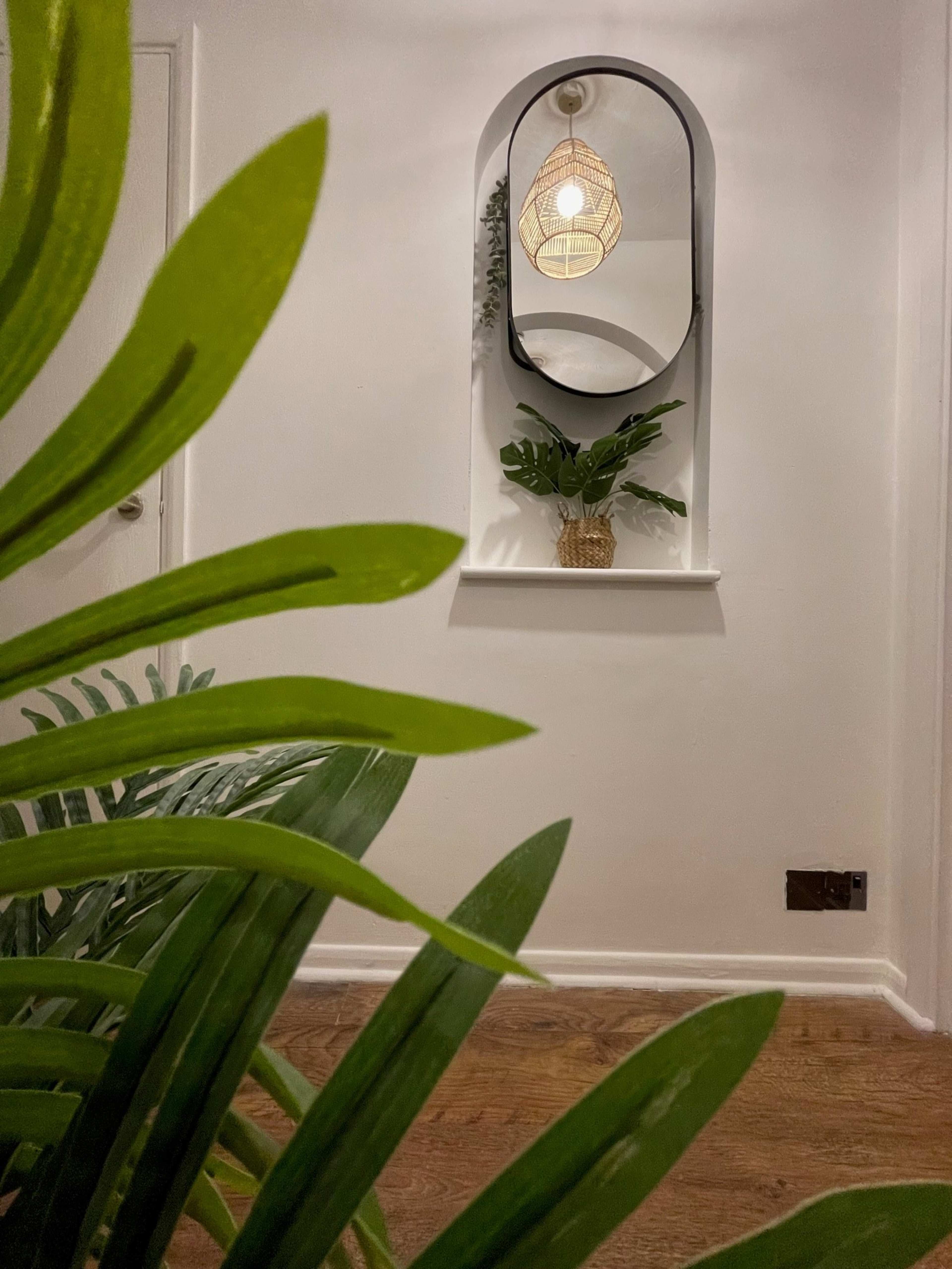 A hallway features a mirror with a woven light fixture above it, surrounded by potted plants on a shelf.