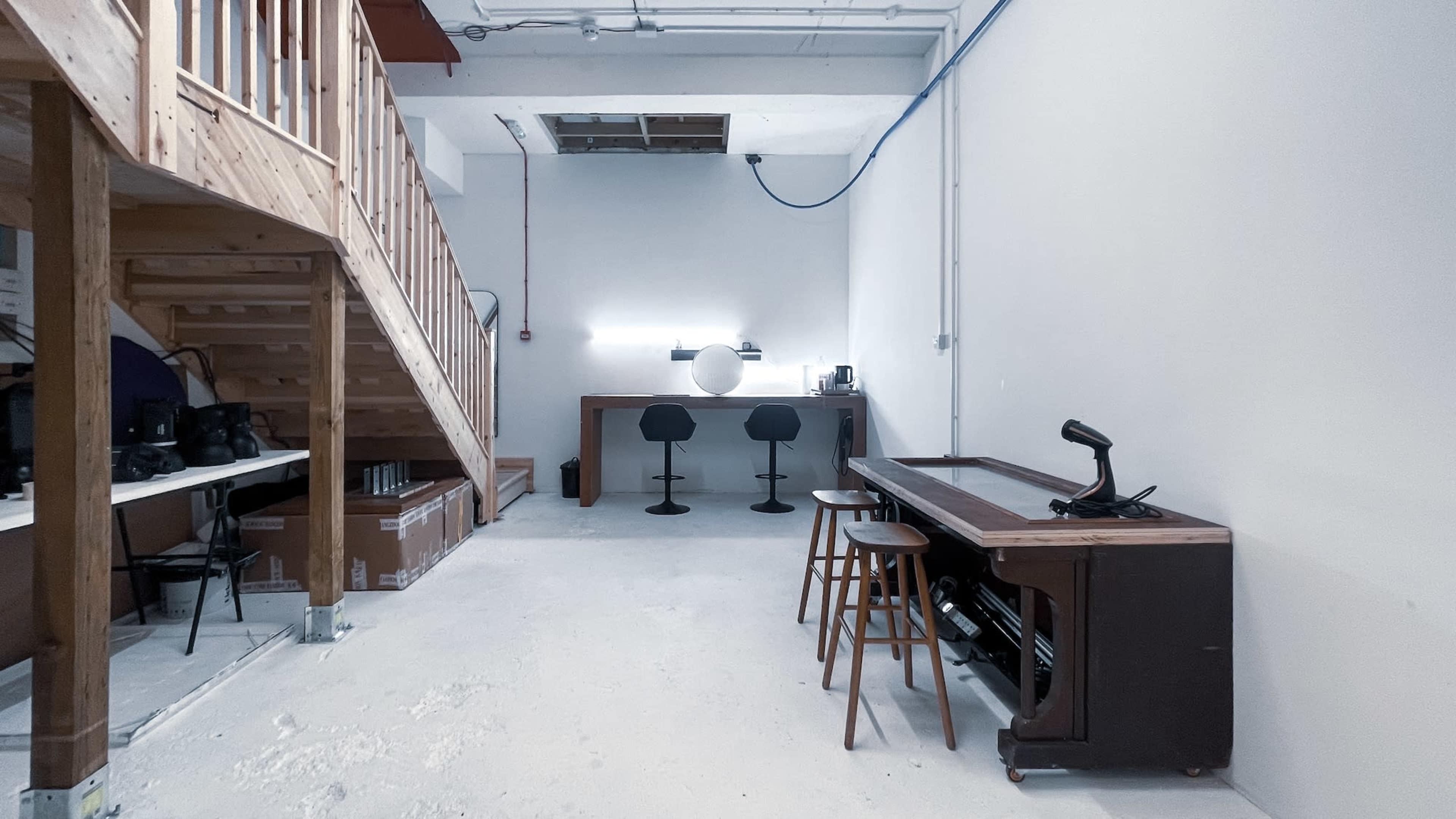 A sparsely furnished studio space features a wooden staircase, a workbench with two stools, and a table with a microphone.