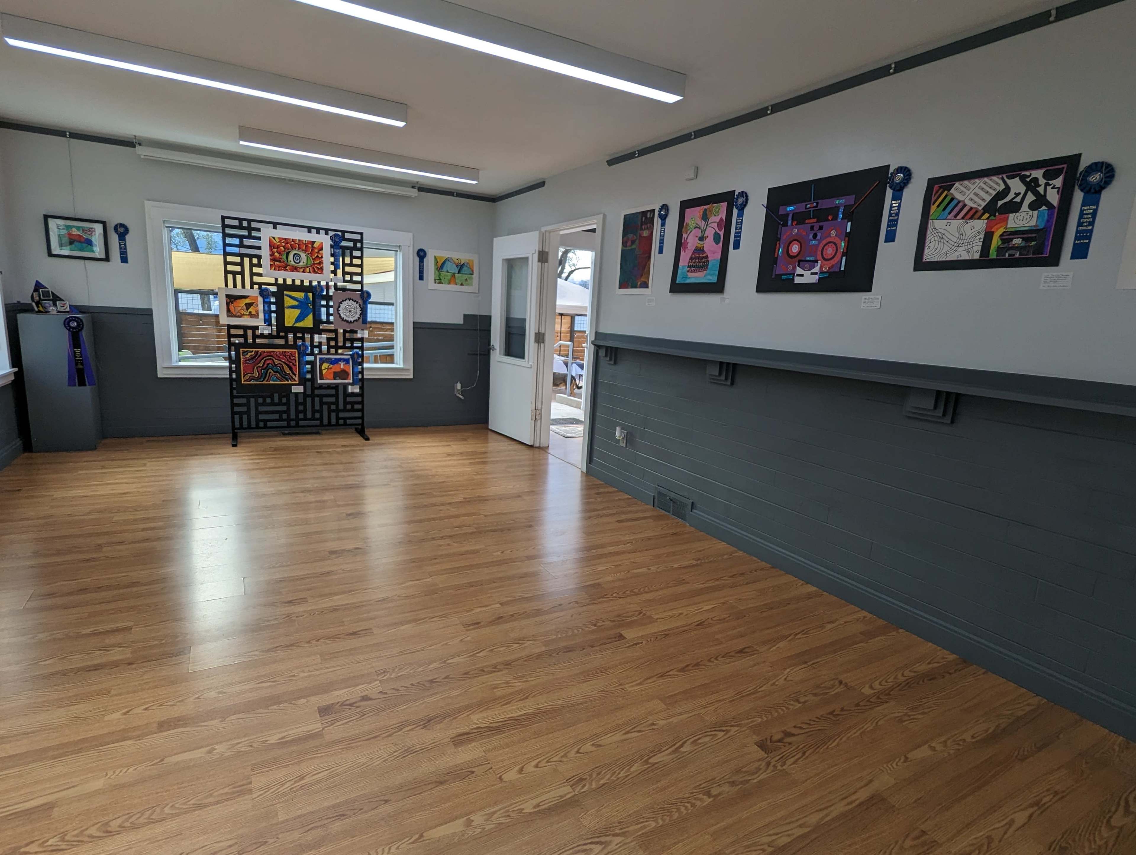 The image shows an empty room with wooden flooring, featuring colorful artwork displayed on the walls and a partition with abstract designs.