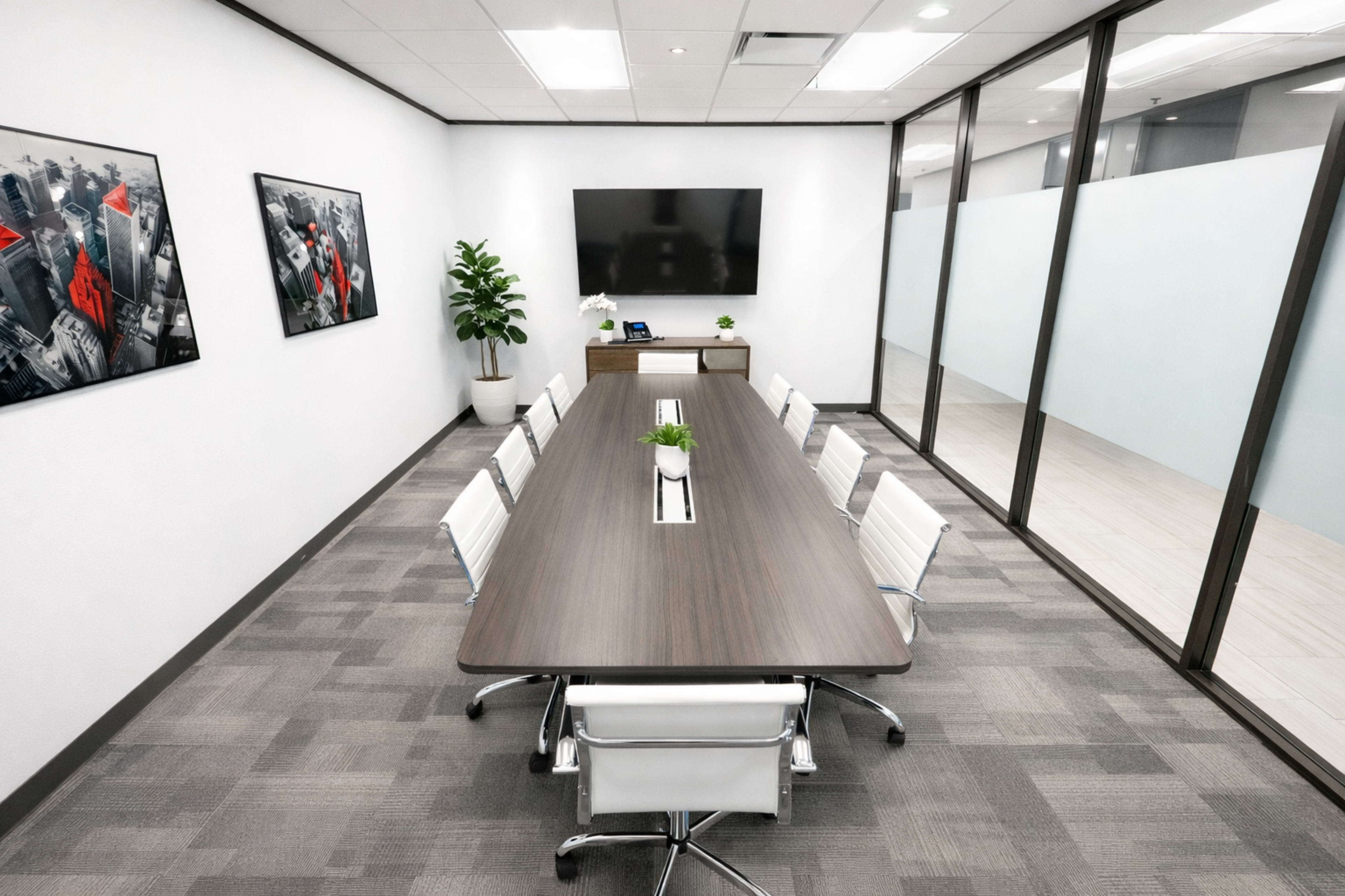 A modern conference room features a long table surrounded by white chairs, with a large screen on the wall and framed artwork on either side.