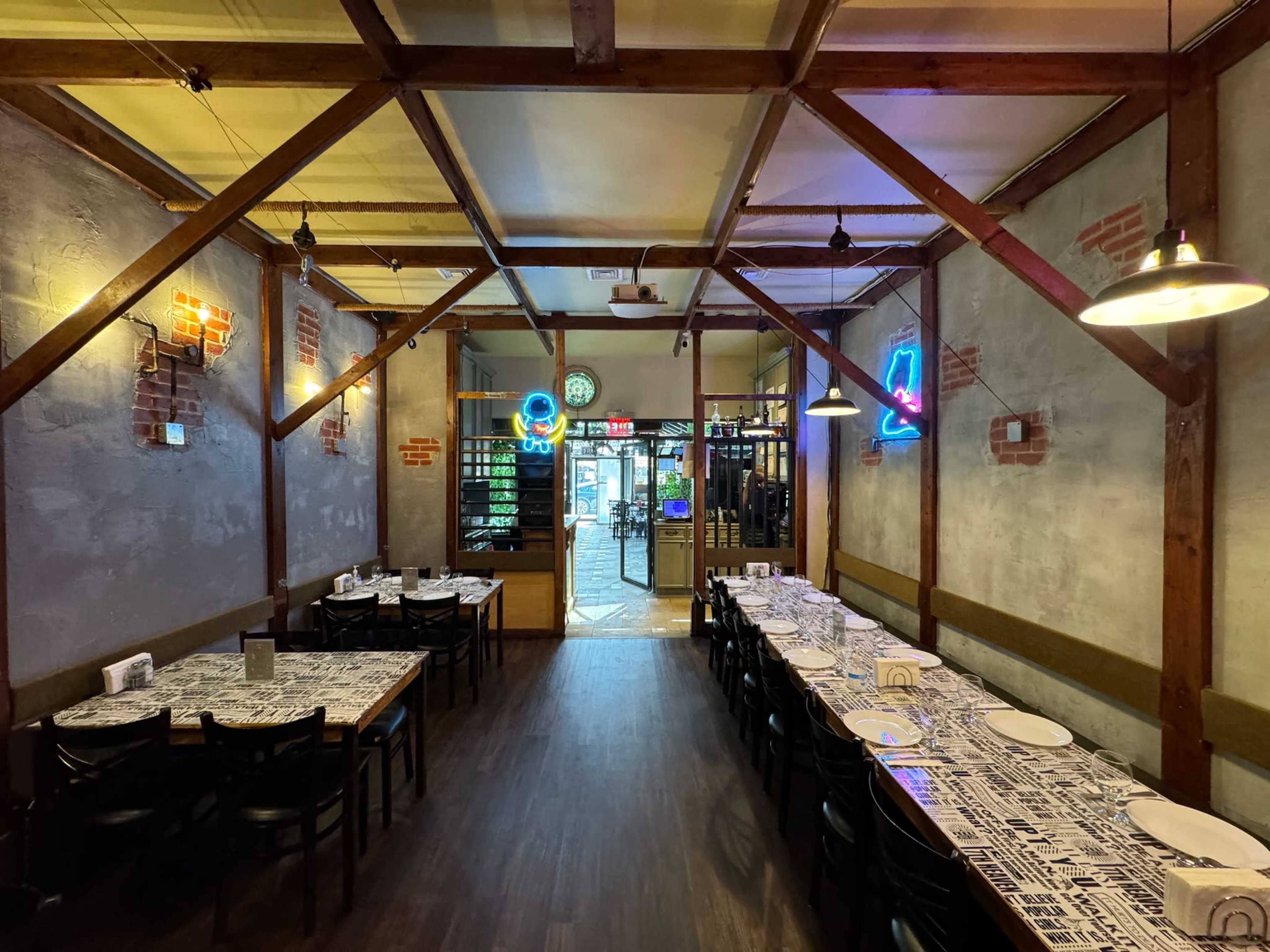 The image shows a rustic restaurant interior featuring a long table set with white plates and wooden beams supporting the ceiling, illuminated by neon signs.