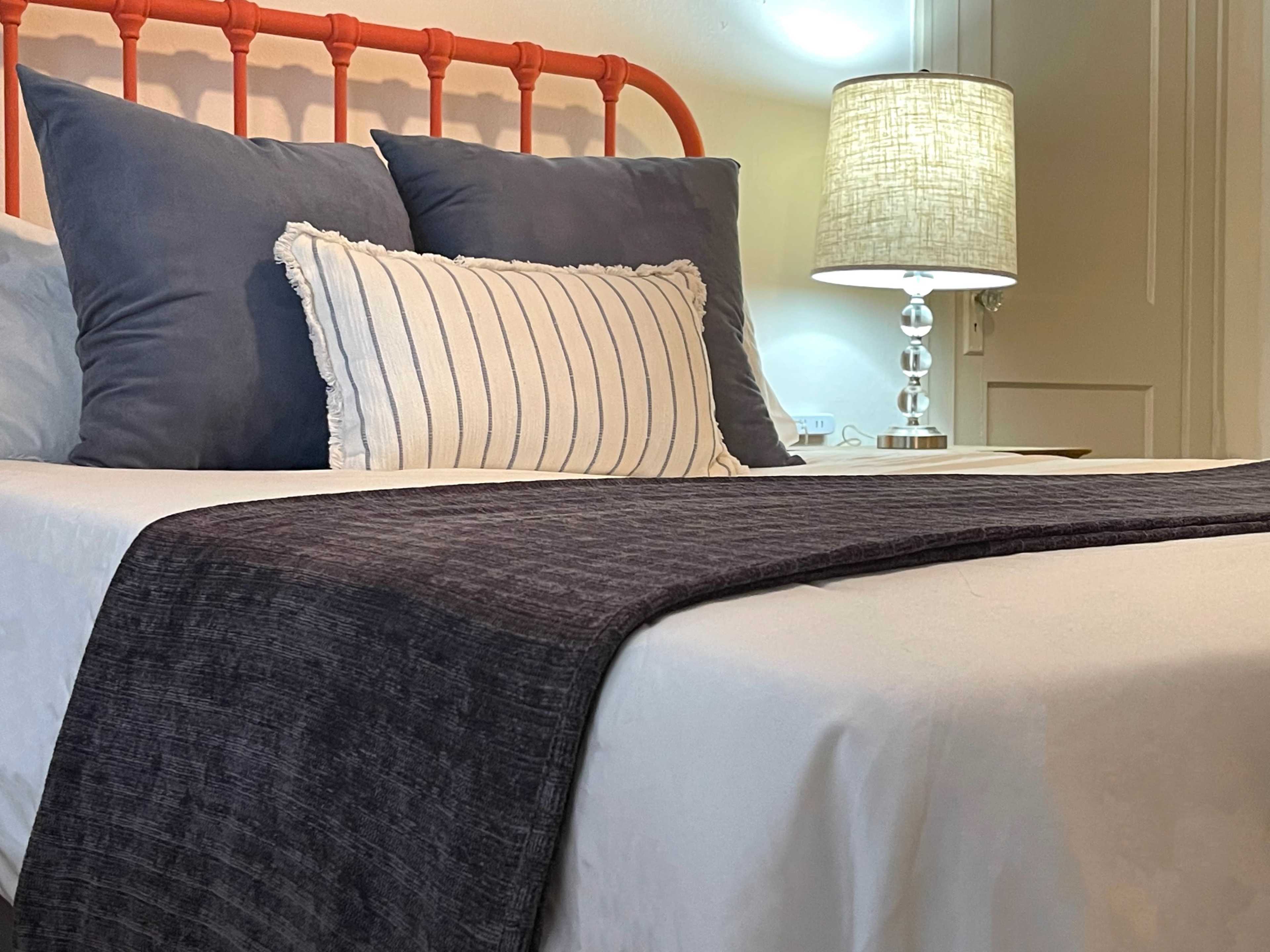 A bed with an orange headboard is adorned with blue and striped pillows, beside a lamp with a textured shade.