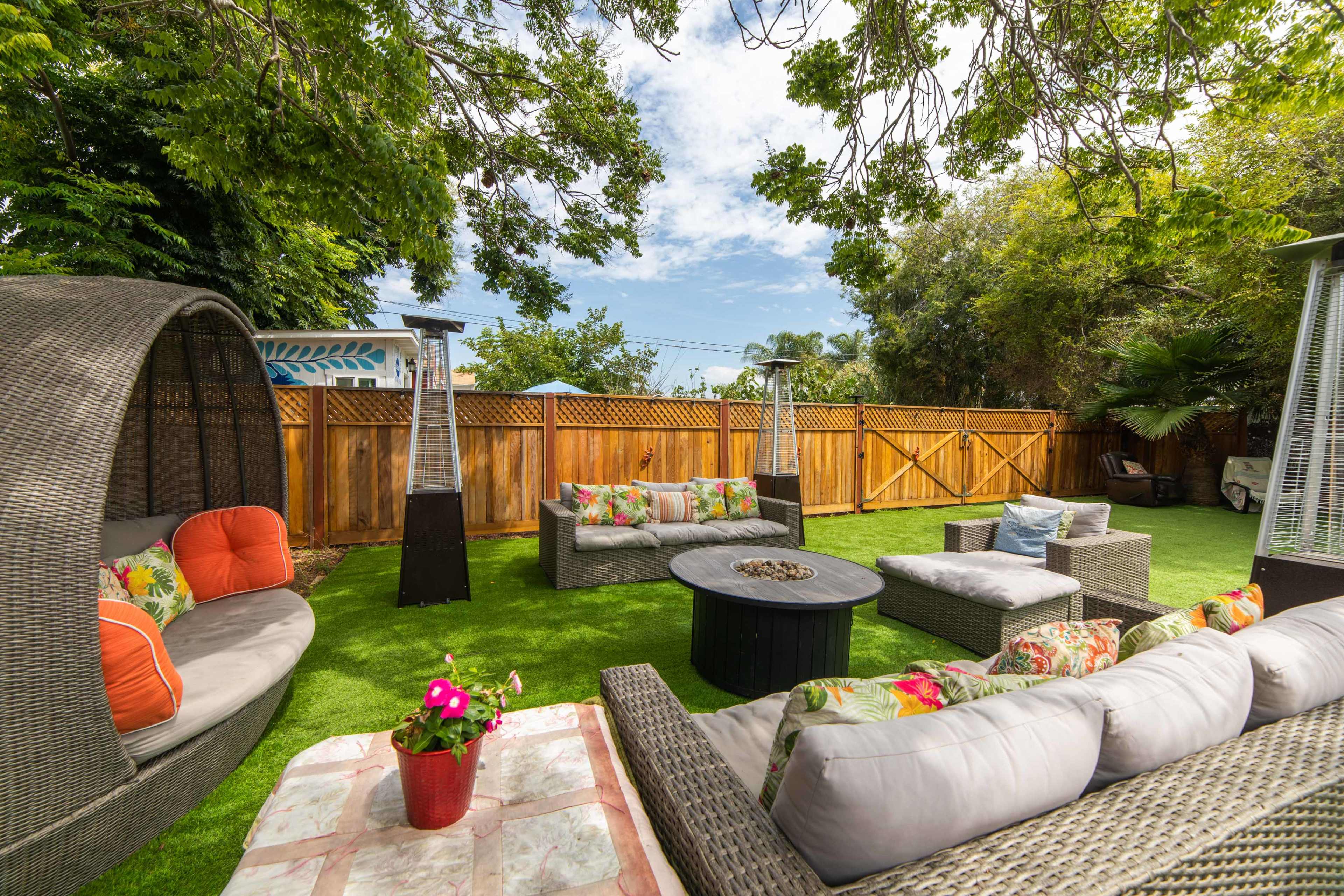 A landscaped backyard features comfortable seating areas, a stone fire pit, and decorative plants under a clear blue sky.