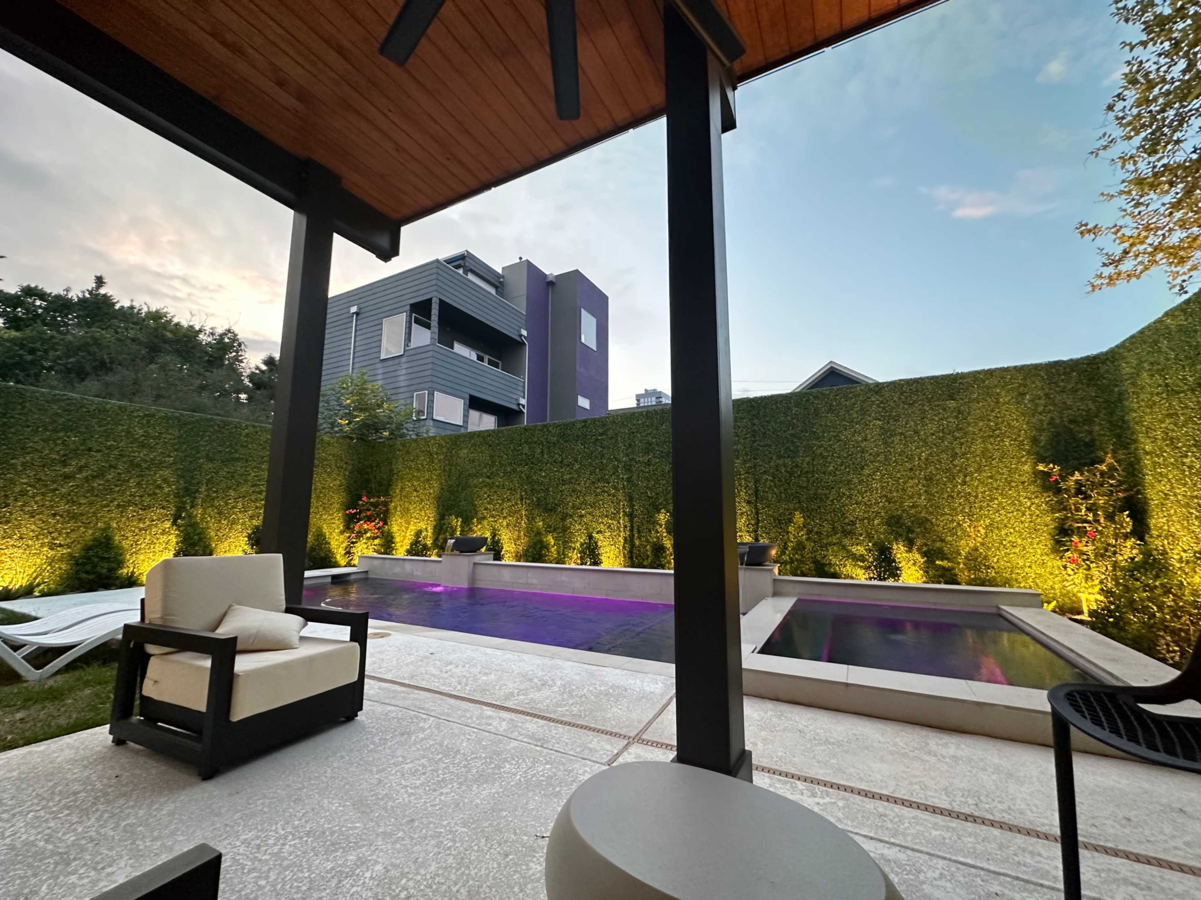 A backyard with a modern pool area surrounded by tall green hedges and outdoor seating beneath a wooden overhang.