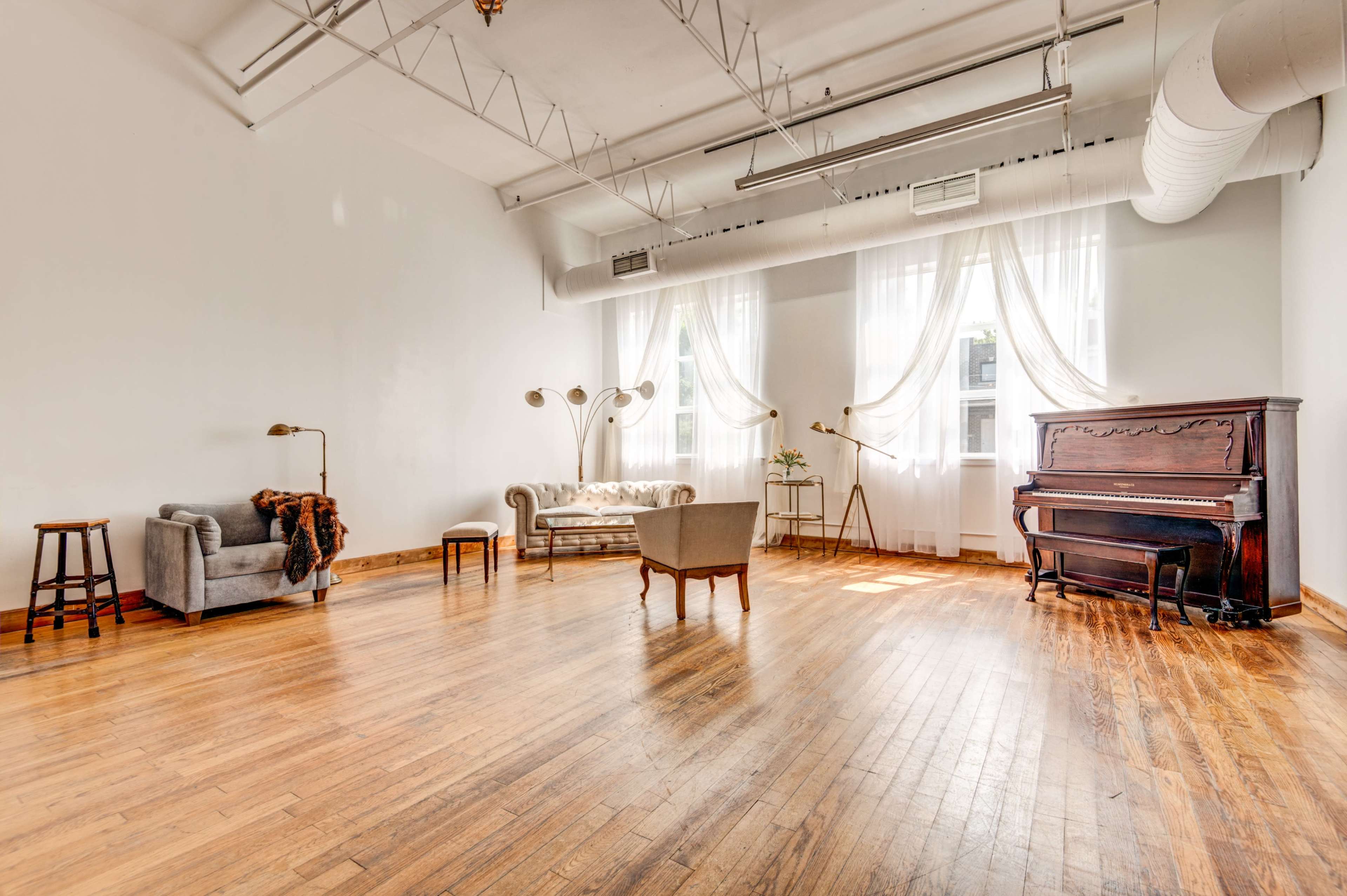 The image shows a spacious, empty room with wooden flooring, large windows covered by sheer curtains, and a mix of vintage and modern furniture, including a piano.