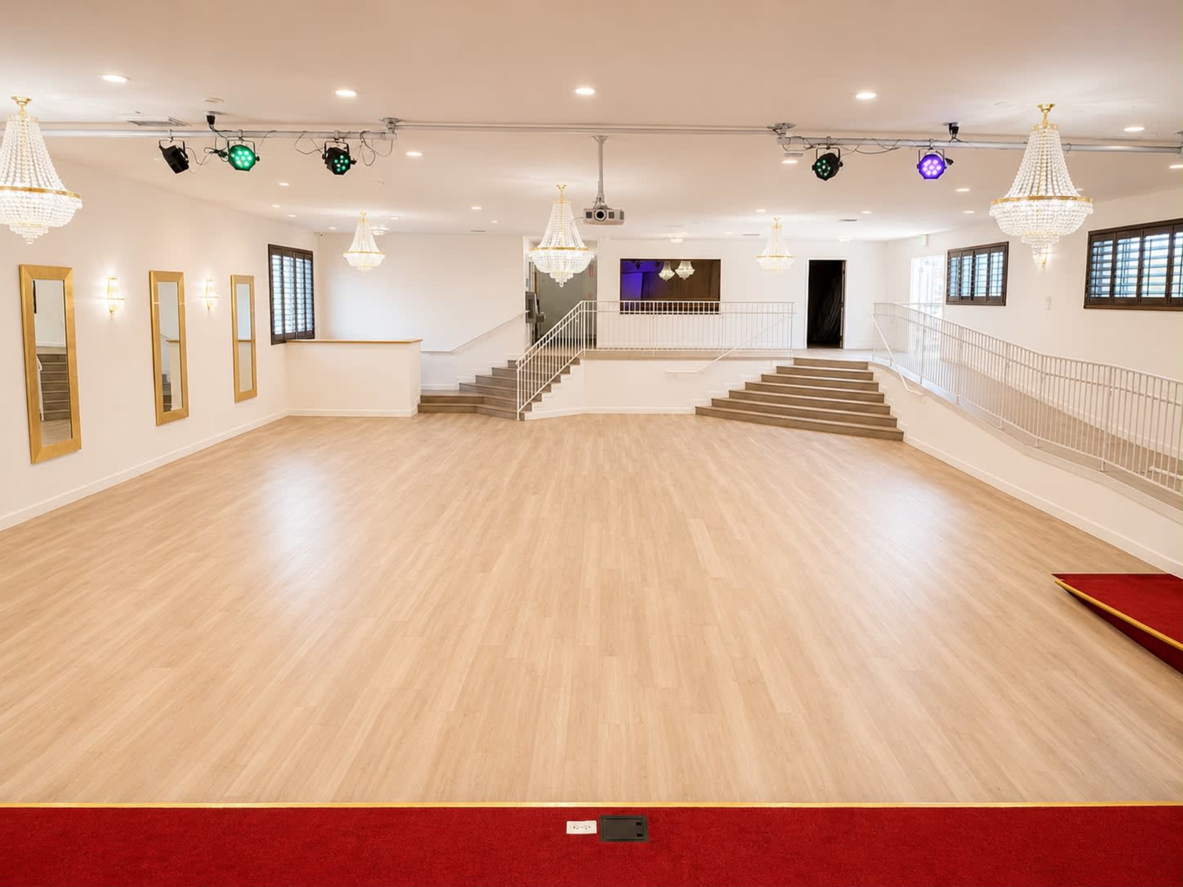 The image shows an empty, spacious event hall with wooden flooring, chandeliers, and a staircase in the background.