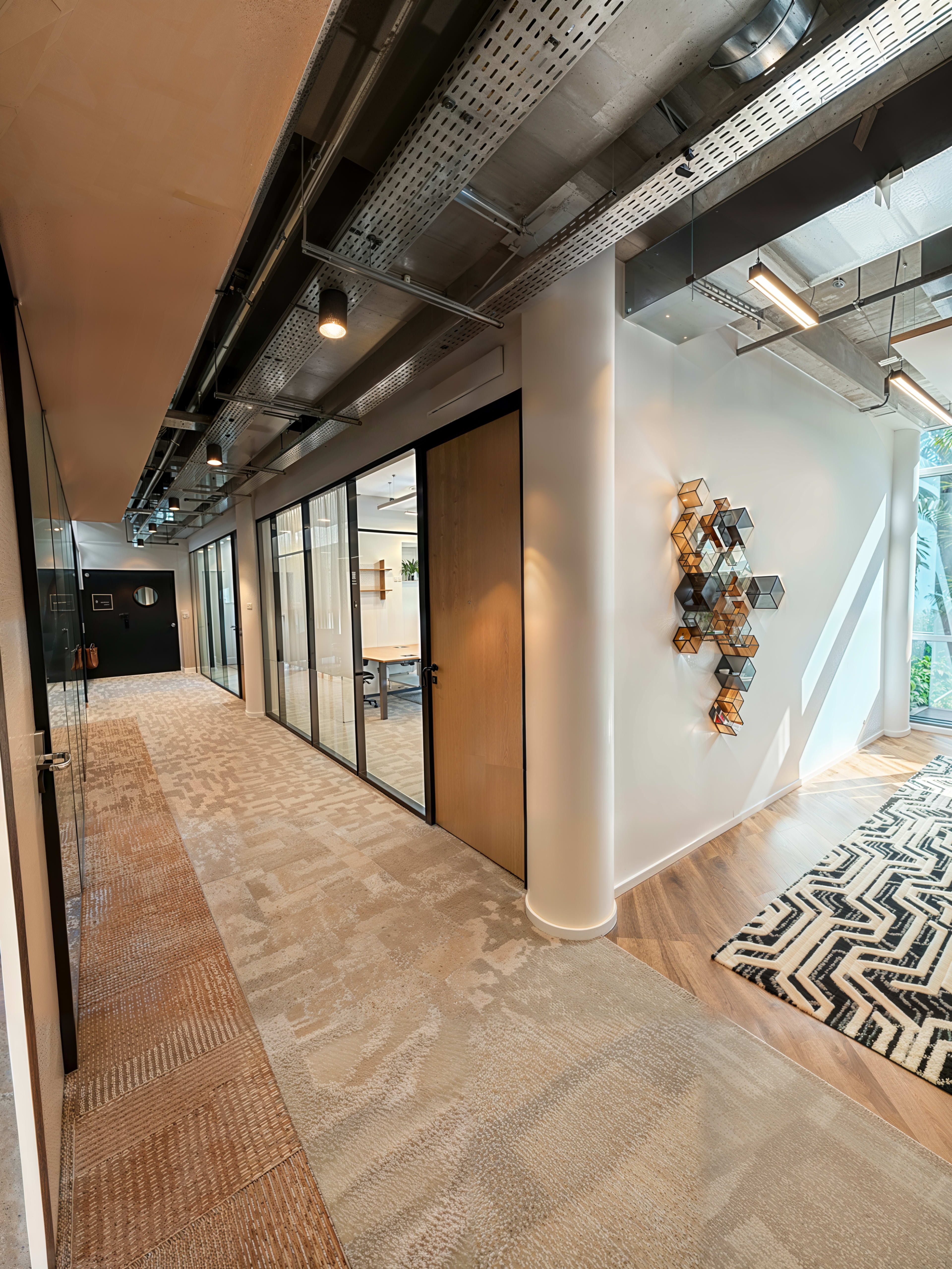 The image depicts a modern office corridor featuring glass-walled meeting rooms, a patterned carpet, and decorative wall art.