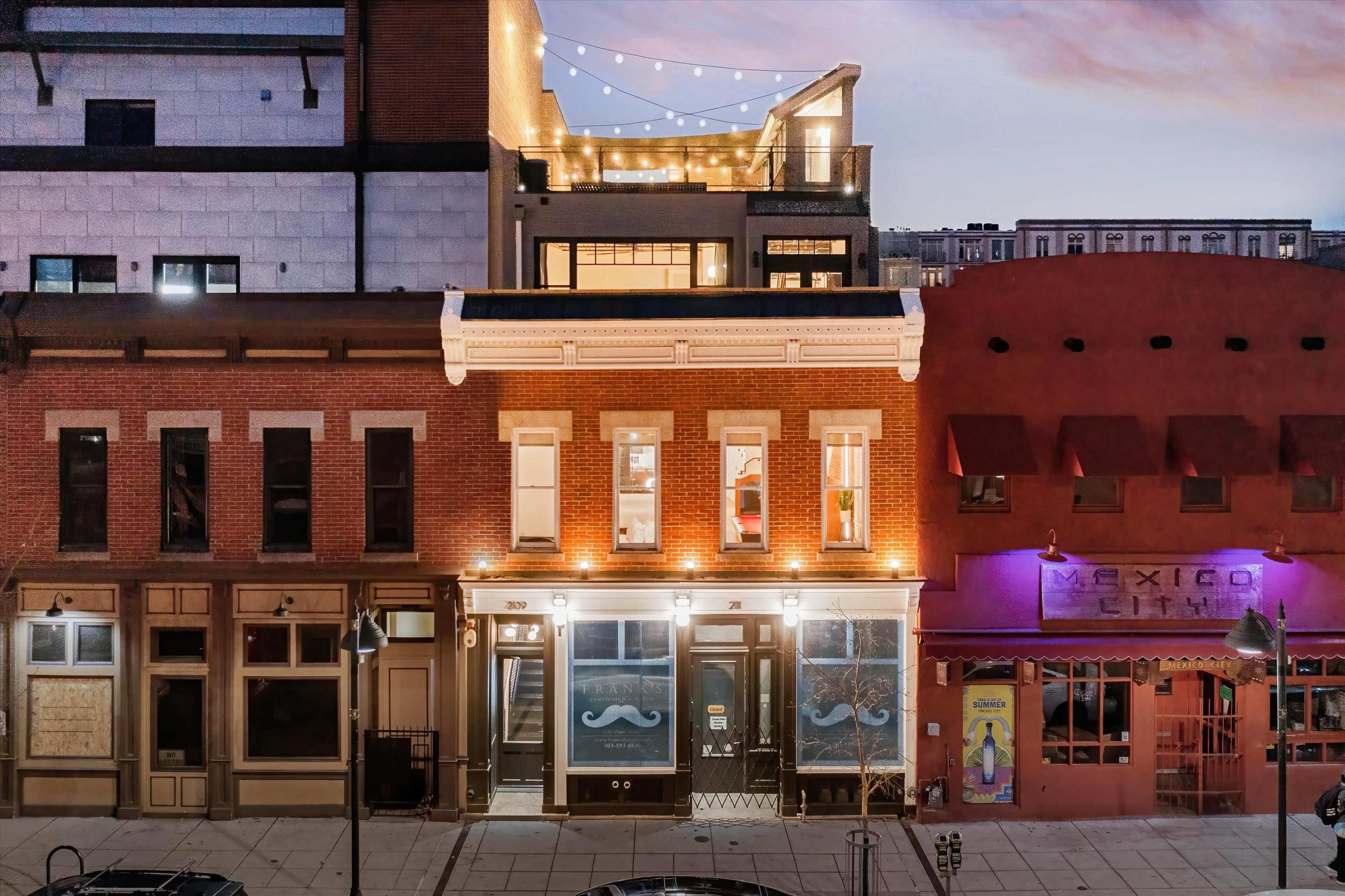 A lively street scene at dusk, featuring well-lit buildings with distinct architectural styles, including a restaurant with a sign displaying a mustache.