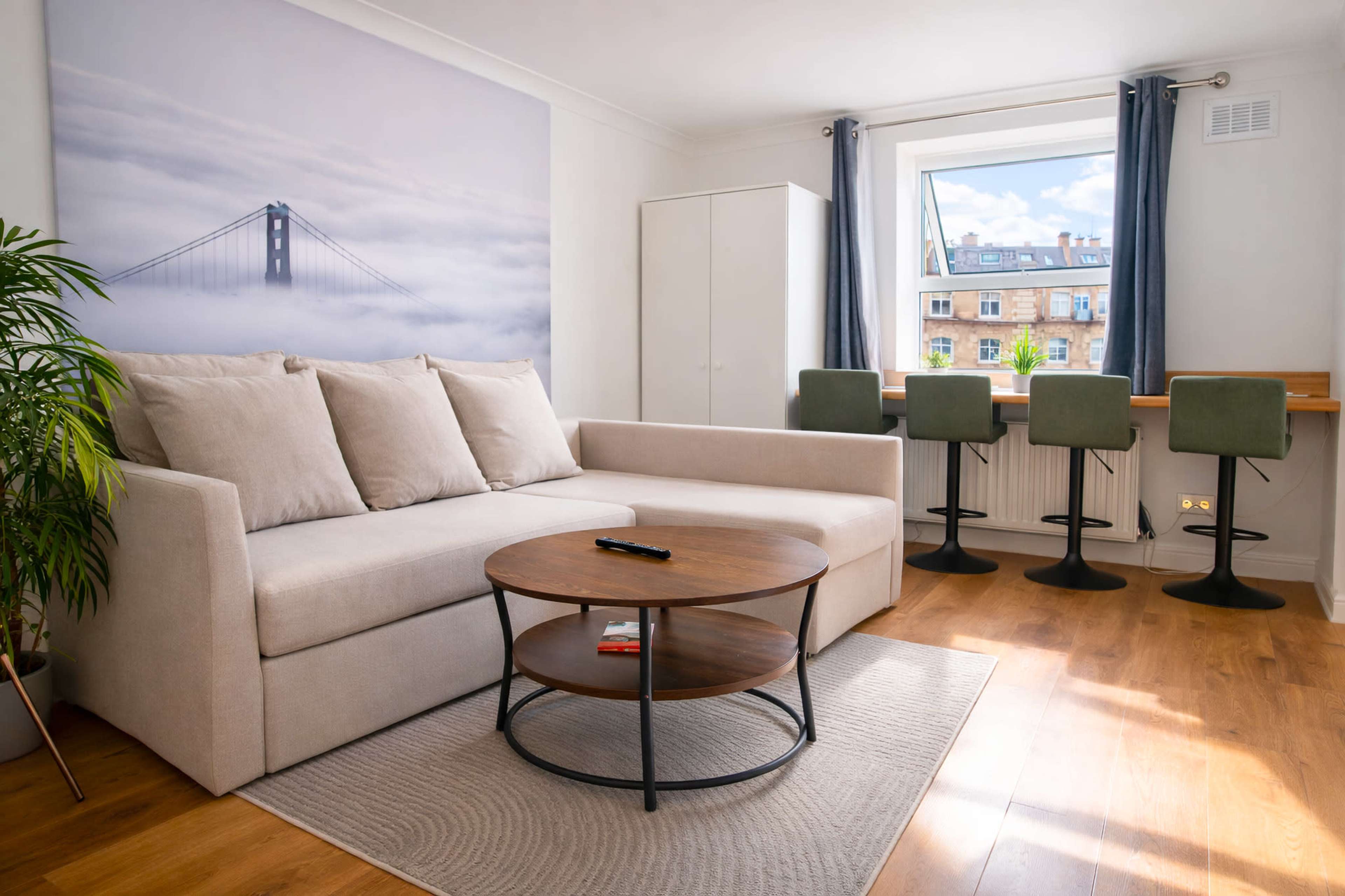 The image shows a modern living room featuring a beige sofa, a round wooden coffee table, and a bar-style seating area with green stools beside a window.