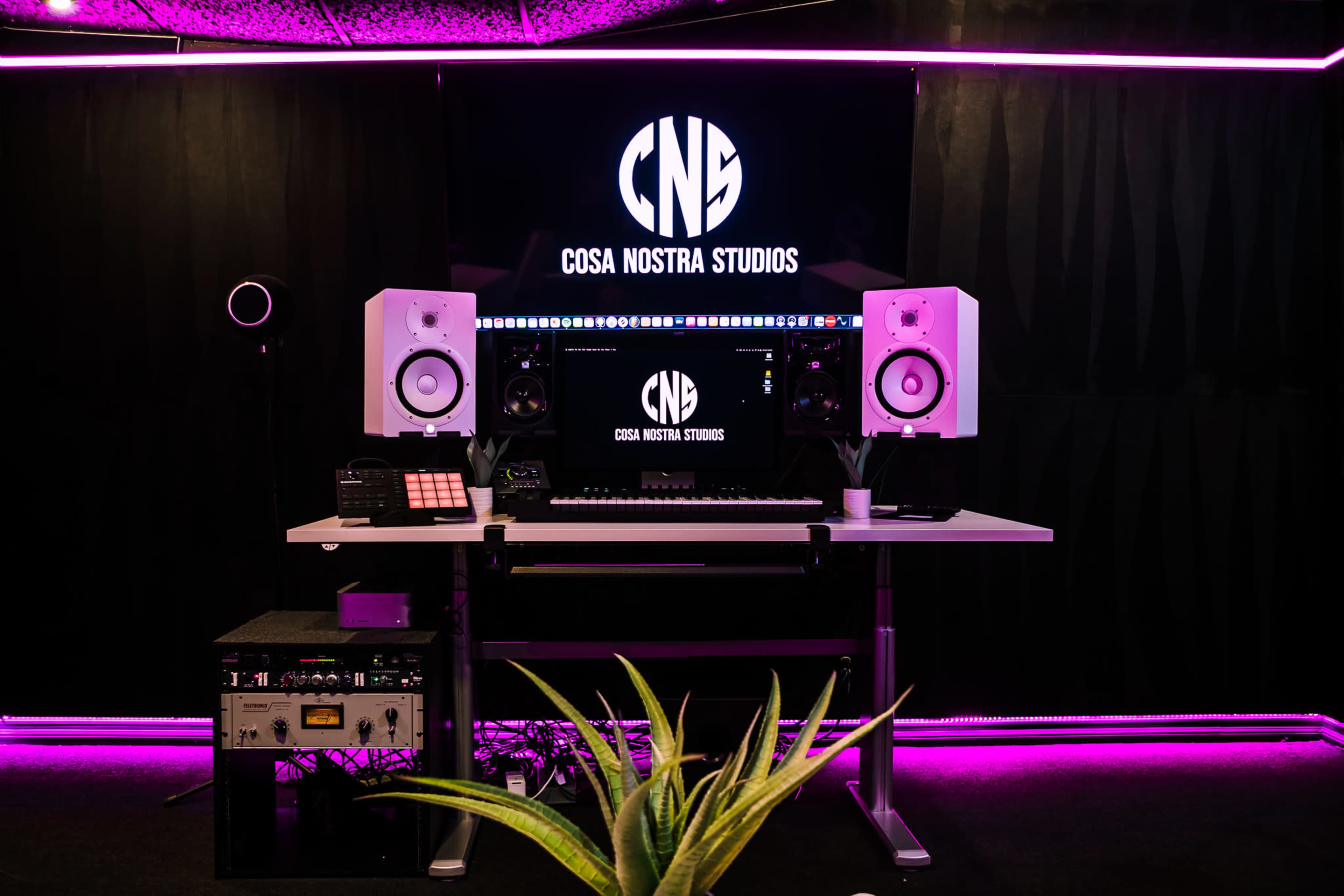 The image shows a music production studio setup with a branded backdrop, monitor speakers, and a control panel.