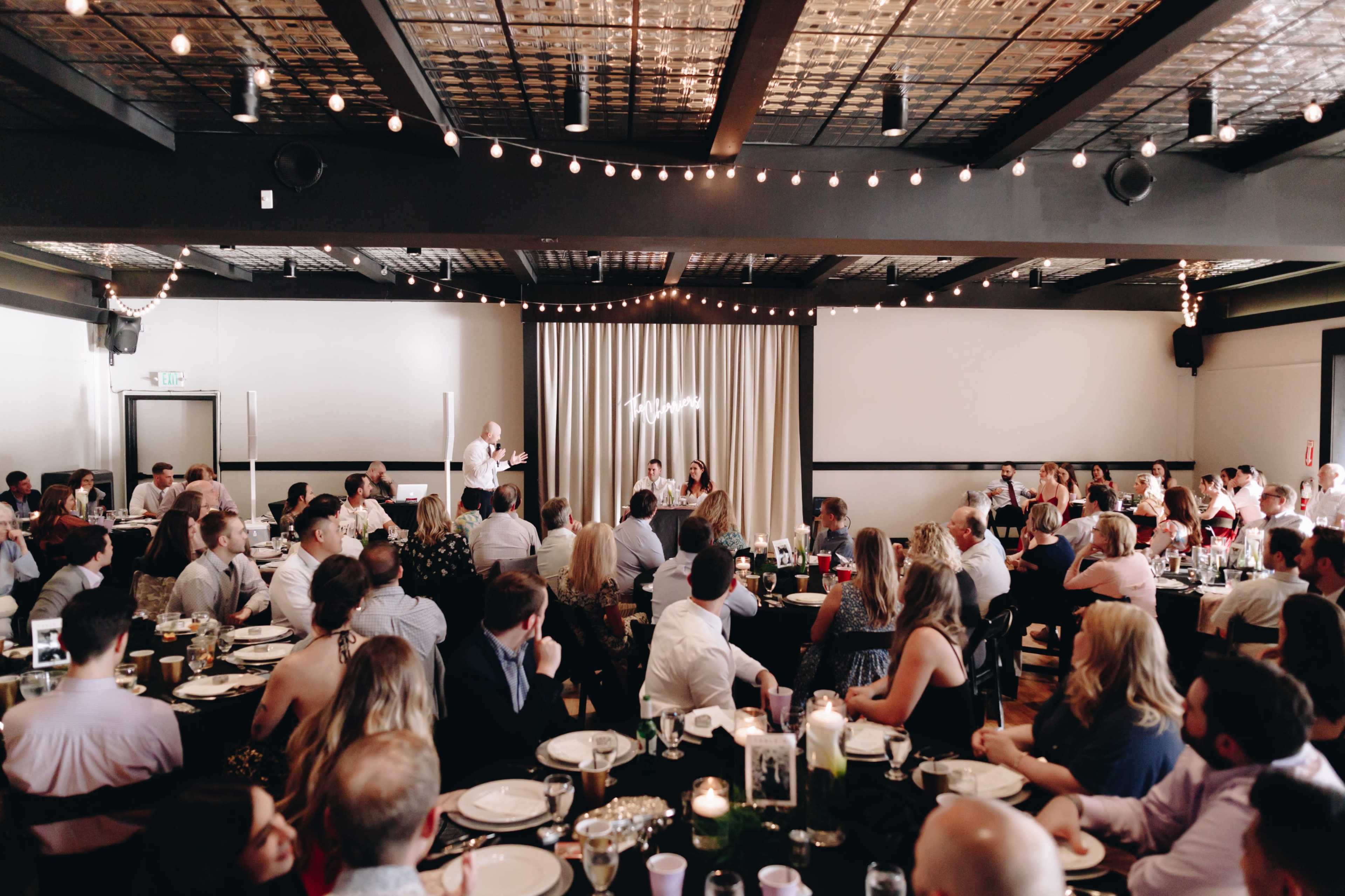 A crowd of guests sits at elegantly set tables in a decorated reception hall, while a speaker addresses the audience near a softly lit backdrop.