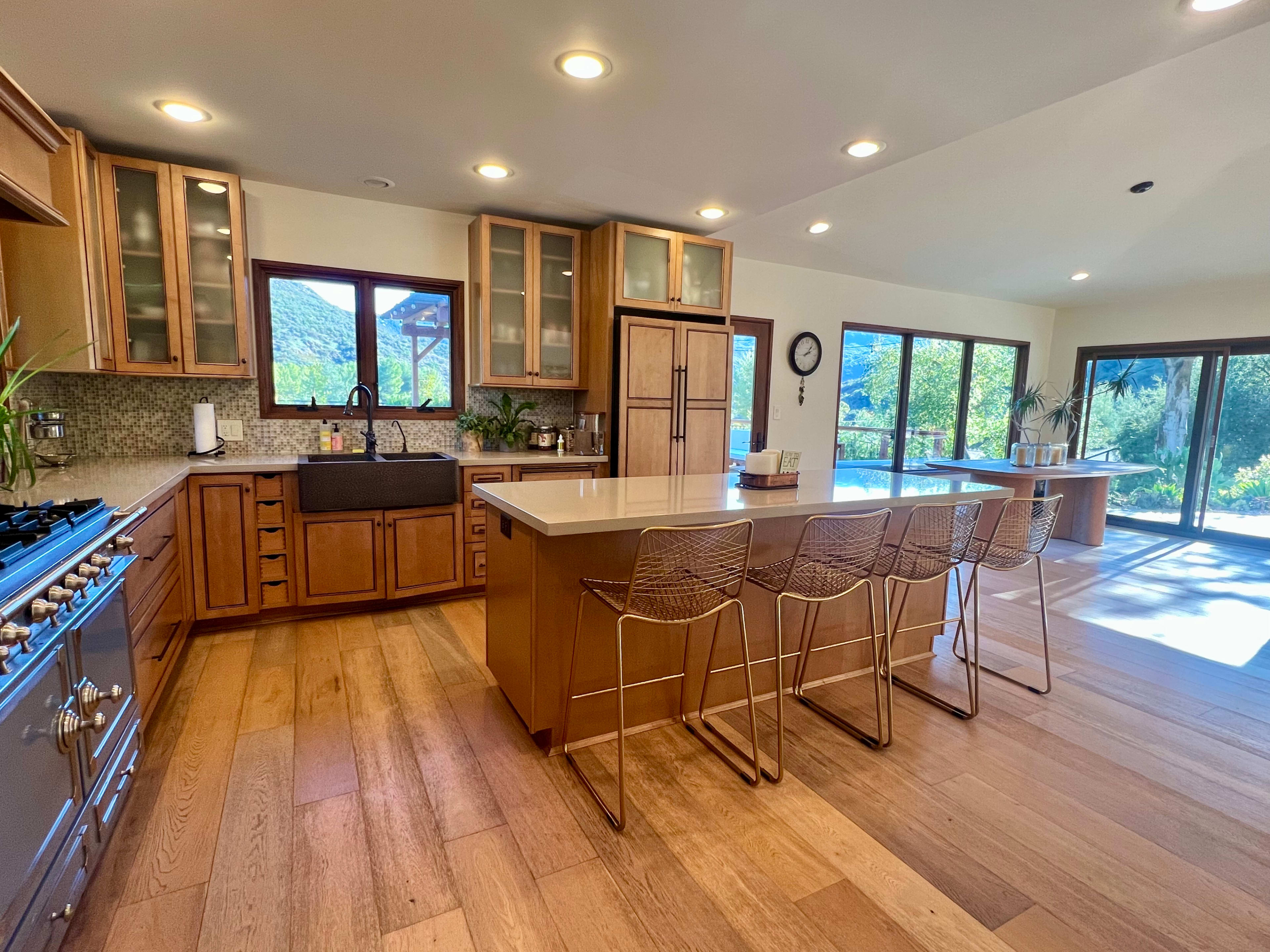The kitchen features a large island with seating, wooden cabinets, and large windows overlooking a natural landscape.