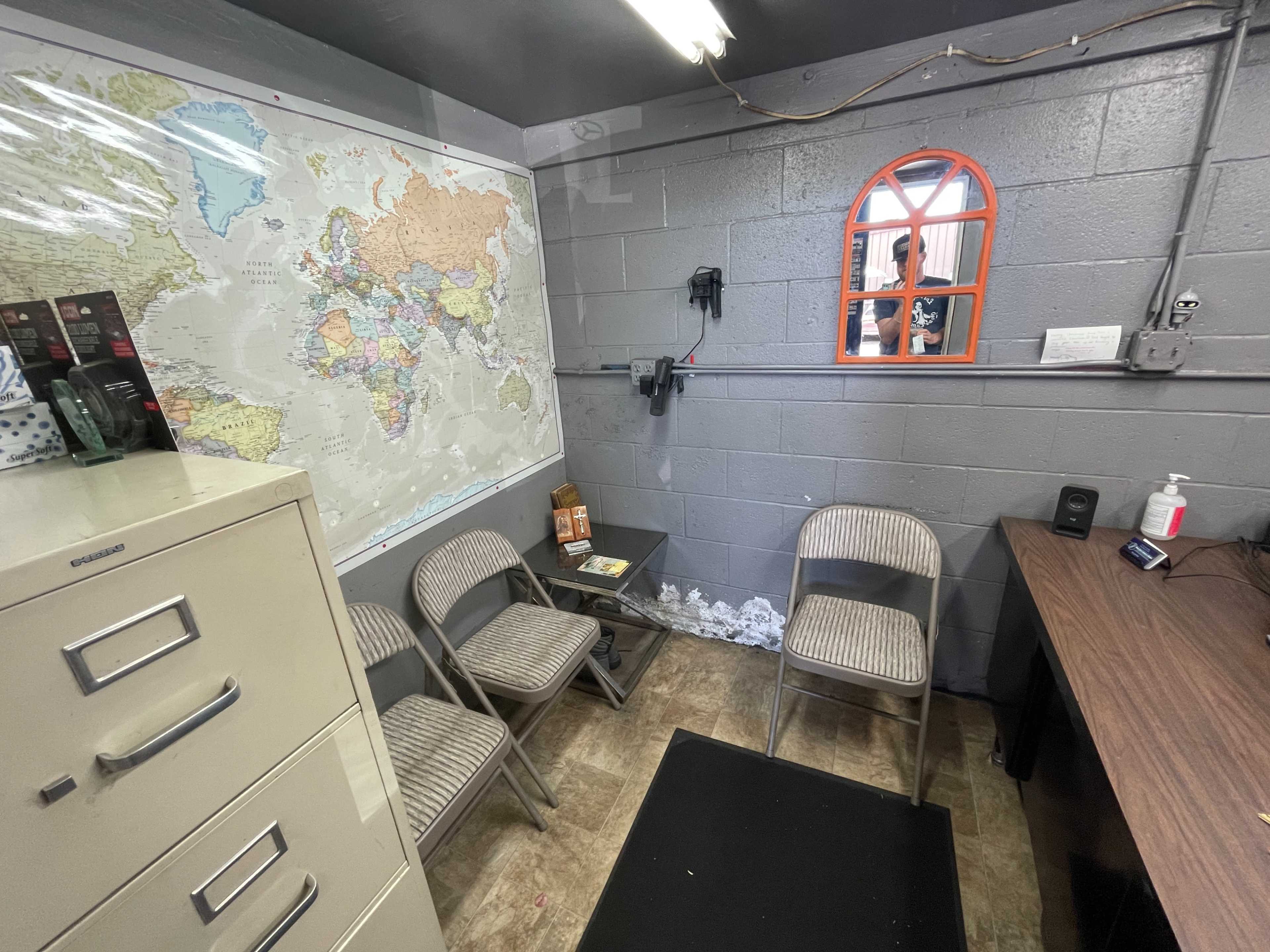 A small room features a world map on the wall, a table surrounded by four chairs, a filing cabinet, and a mirror above a desk.
