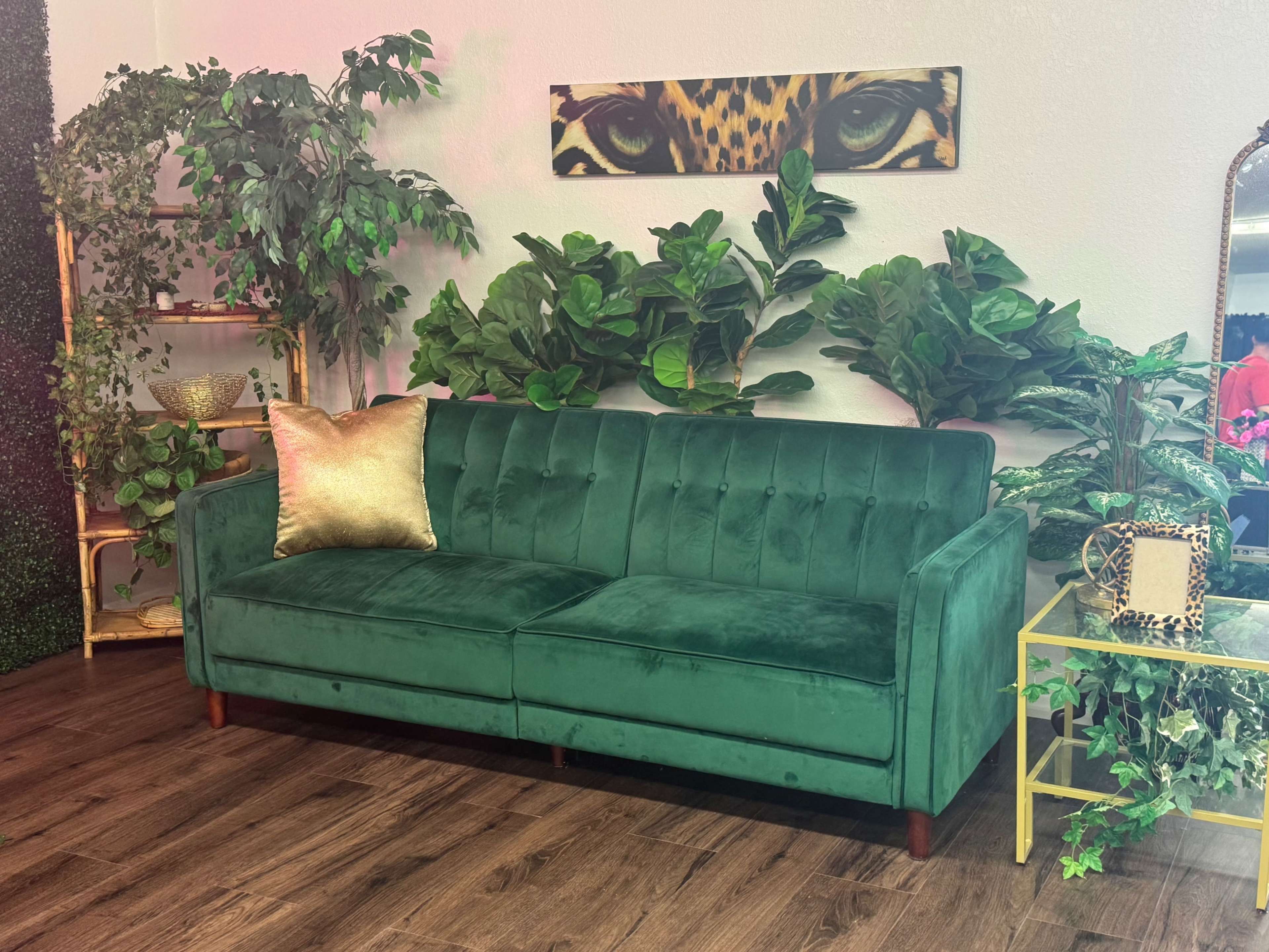 The image shows a green velvet sofa with a gold accent pillow, positioned among various indoor plants and decorated with a gold side table.