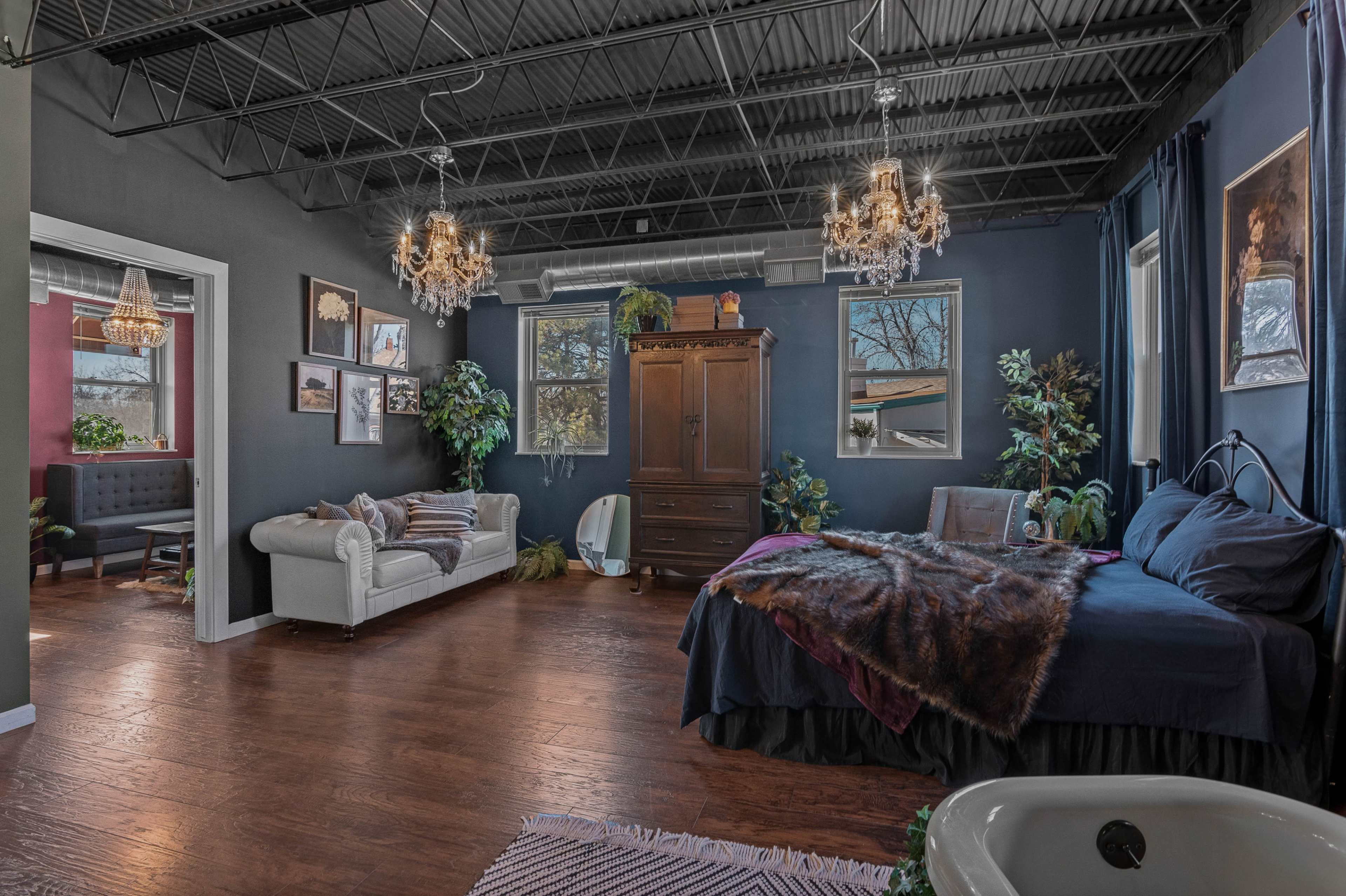The image shows a bedroom with a dark-painted wall, a bed with a fur throw, a white couch, wooden flooring, and various decorative plants and chandeliers.