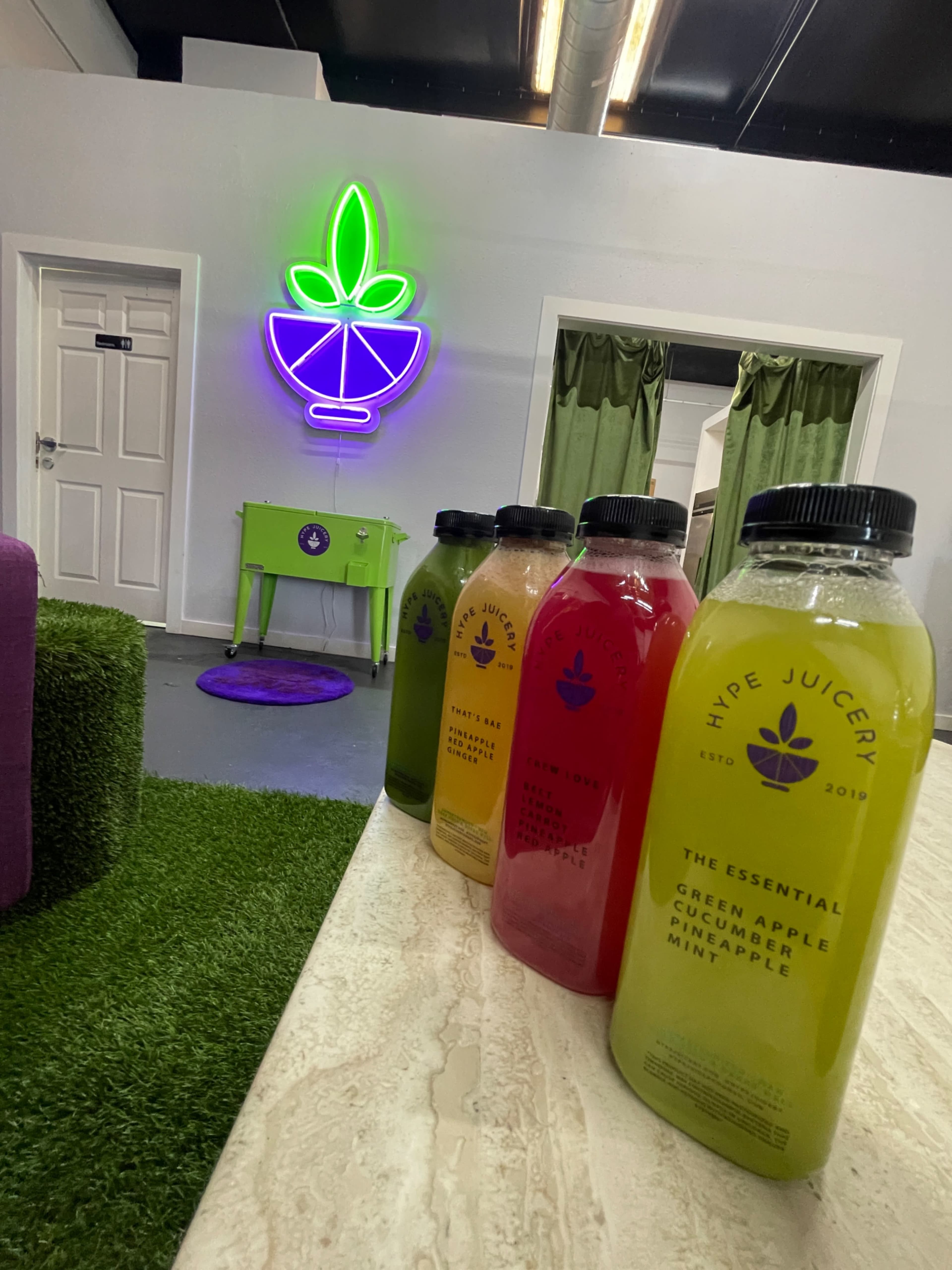 Several colorful juice bottles lined up on a countertop in a juice bar with a neon sign of a leaf-shaped logo on the wall.